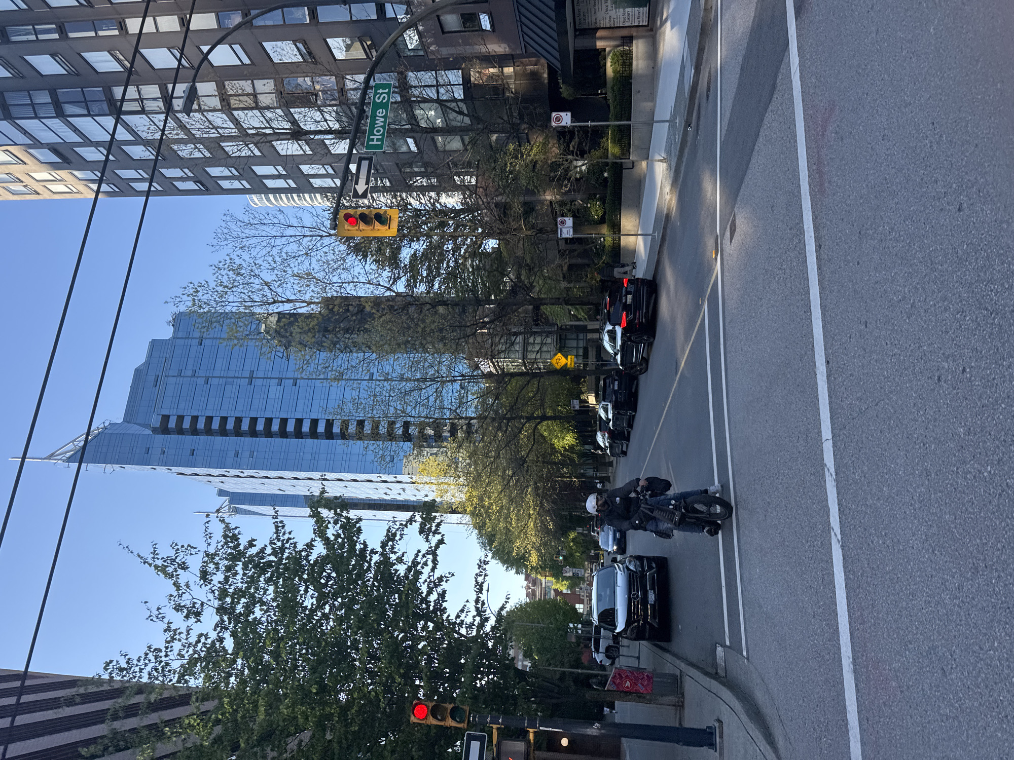 This image depicts an urban street scene in Vancouver, Canada, under bright daylight with a clear blue sky. The view is from a slightly elevated perspective, looking down a multi-lane city street.
The street is identified by a green sign as "Howe St," accompanied by a left-turn arrow. A traffic light showing a red signal is prominently visible on a pole, suggesting that vehicles are stopped. On the street, a person wearing a white helmet and dark clothing is seated on a motorcycle in the foreground, next to a dark-colored car. Several other cars, primarily black SUVs, are visible further down the street, also appearing to be stopped.
The street is bordered by various buildings. On the left, modern skyscrapers with extensive glass facades reflect the blue sky and other structures. On the right, older buildings with numerous windows rise above street level. Green trees with lush foliage line both sides of the street, providing natural elements within the urban setting.
Visible street furniture includes power lines or cables stretching across the scene, additional street signs (one indicating "No Stopping"), and a fire hydrant on the sidewalk. The strong sunlight casts clear shadows, indicating a sunny day, likely mid-morning or afternoon. White lines are clearly marked on the asphalt road, delineating lanes and pedestrian crossings. The overall scene portrays a bustling city environment.