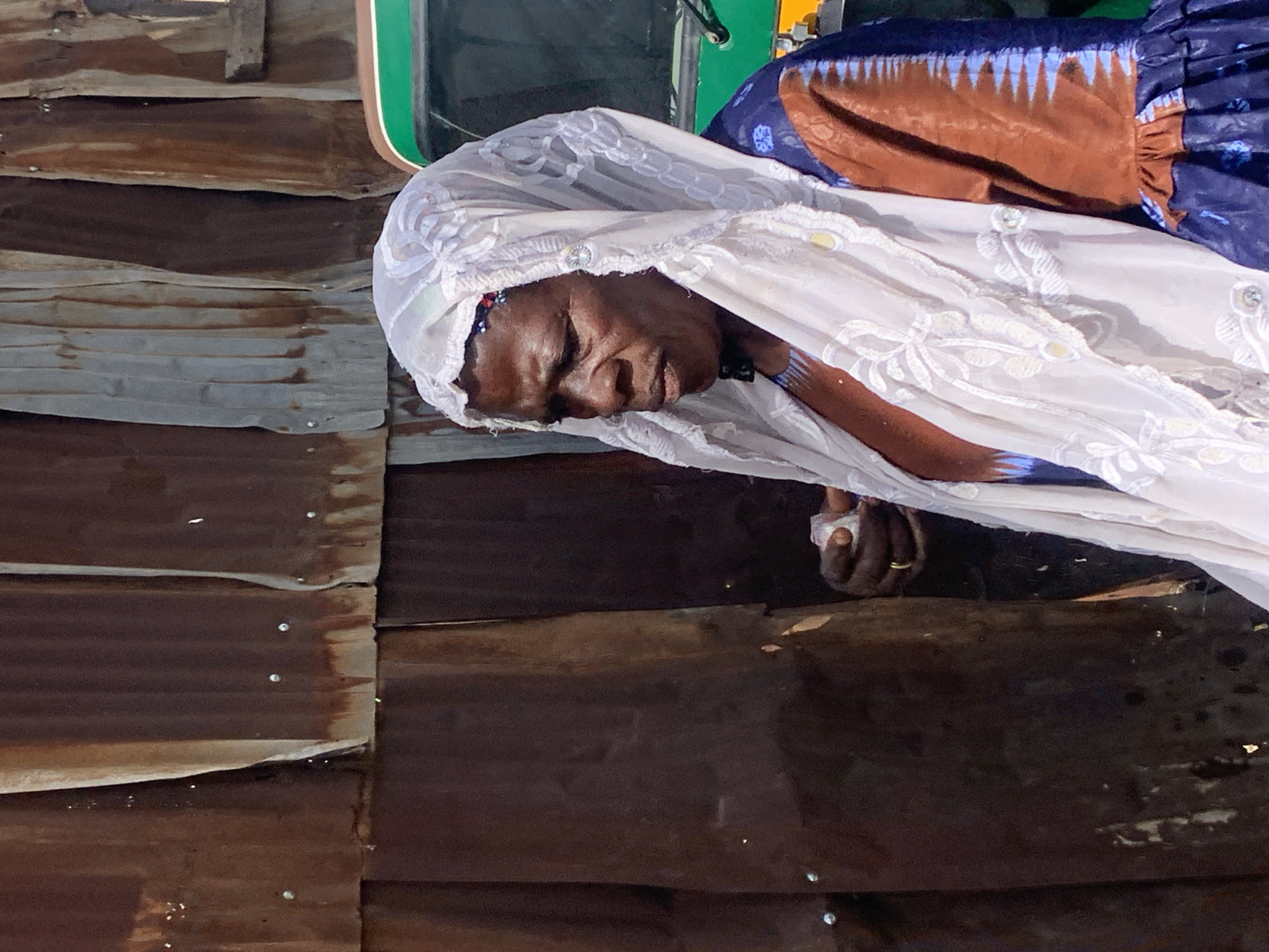 The image shows a close-up of an elderly Black woman wearing a white headscarf with intricate lace detailing. Her eyes are closed, and her head is tilted downwards, suggesting a moment of rest or contemplation. She is wearing a patterned blue and brown garment beneath the headscarf. Her hands are visible, one of which is clasped and has a gold ring on one finger. The background consists of corrugated metal panels, likely forming a wall or structure, with a wooden element visible at the top. The overall lighting suggests it is daytime. The scene appears to be outdoors, given the rustic nature of the background materials. The woman's facial expression is serene.