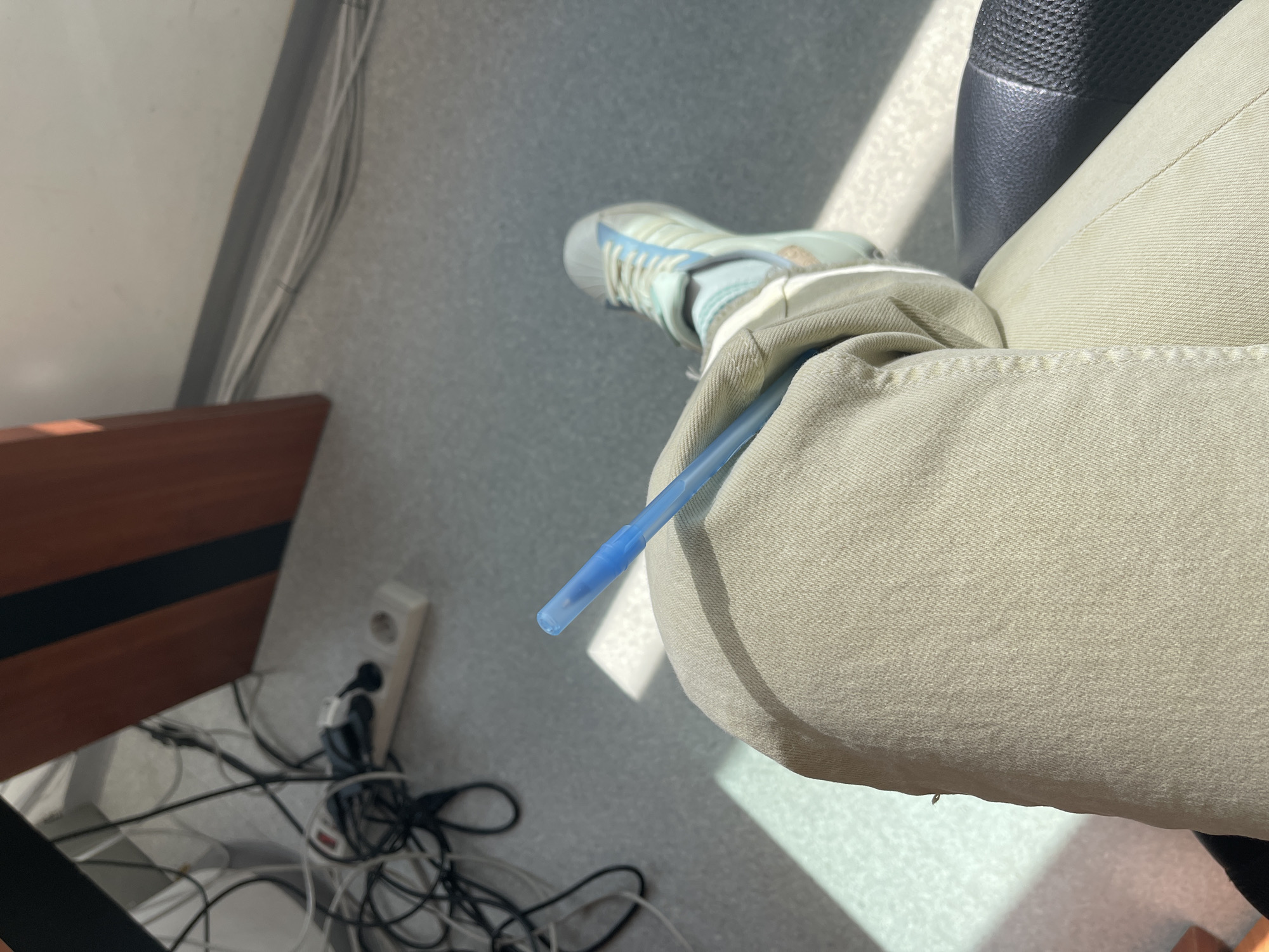 The scene is an indoor shot, likely taken in an office or workspace. A person's leg, clad in khaki-colored pants, is crossed at the knee. A blue ballpoint pen is tucked between the leg and the upper thigh. The person is wearing a light blue and white sneaker. Below the leg, on the floor, are tangled black and white cables, which connect to a white power outlet. A section of dark wood furniture is partially visible in the left background. The floor is a light gray, suggesting a commercial or institutional setting. The light seems to be natural, entering from the side. The location in Sofia, Bulgaria, cannot be determined from the visual data provided.