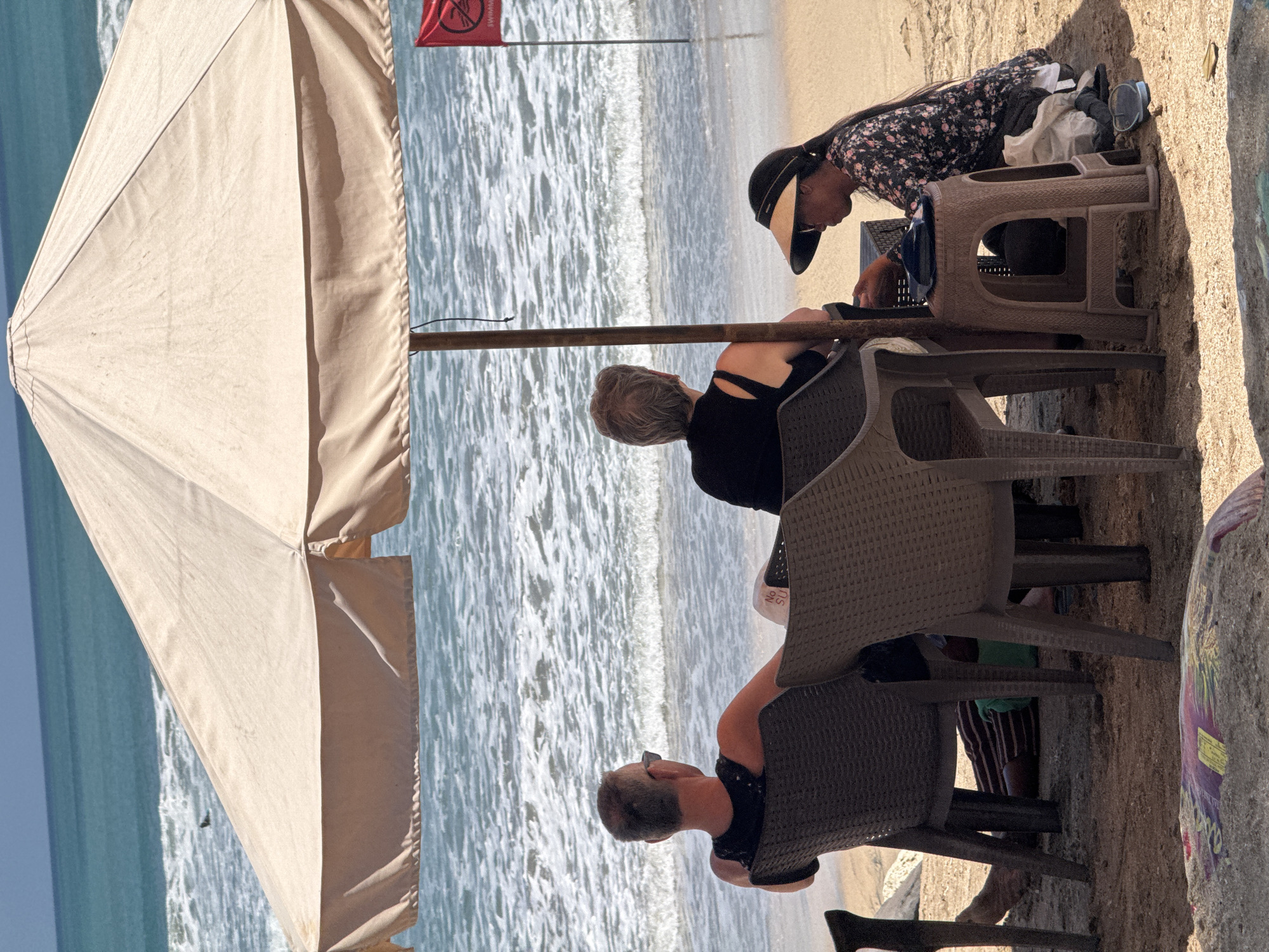The image depicts a beach scene in Kuta, Indonesia, on a sunny day with clear skies and calm ocean waters. Three individuals are present. In the foreground, a man and a woman are seated in beach chairs under a large, light-colored umbrella, facing the ocean. The man, with short grey hair and sunglasses, wears a black tank top. The woman, with short grey hair, is also seated, wearing a black top. To their right, a young woman is crouched on the sand, wearing a dark floral-patterned shirt, a sun visor, and holding a small stool. She is looking down at something in her hands.
The beach is sandy, with some debris scattered on the ground, including a colorful mat. The ocean exhibits gentle waves and a few people can be seen in the water in the distance. A red flag with a white symbol is planted in the sand behind the seated individuals, indicating a no-swimming zone. The overall atmosphere is relaxed and typical of a beach resort area.