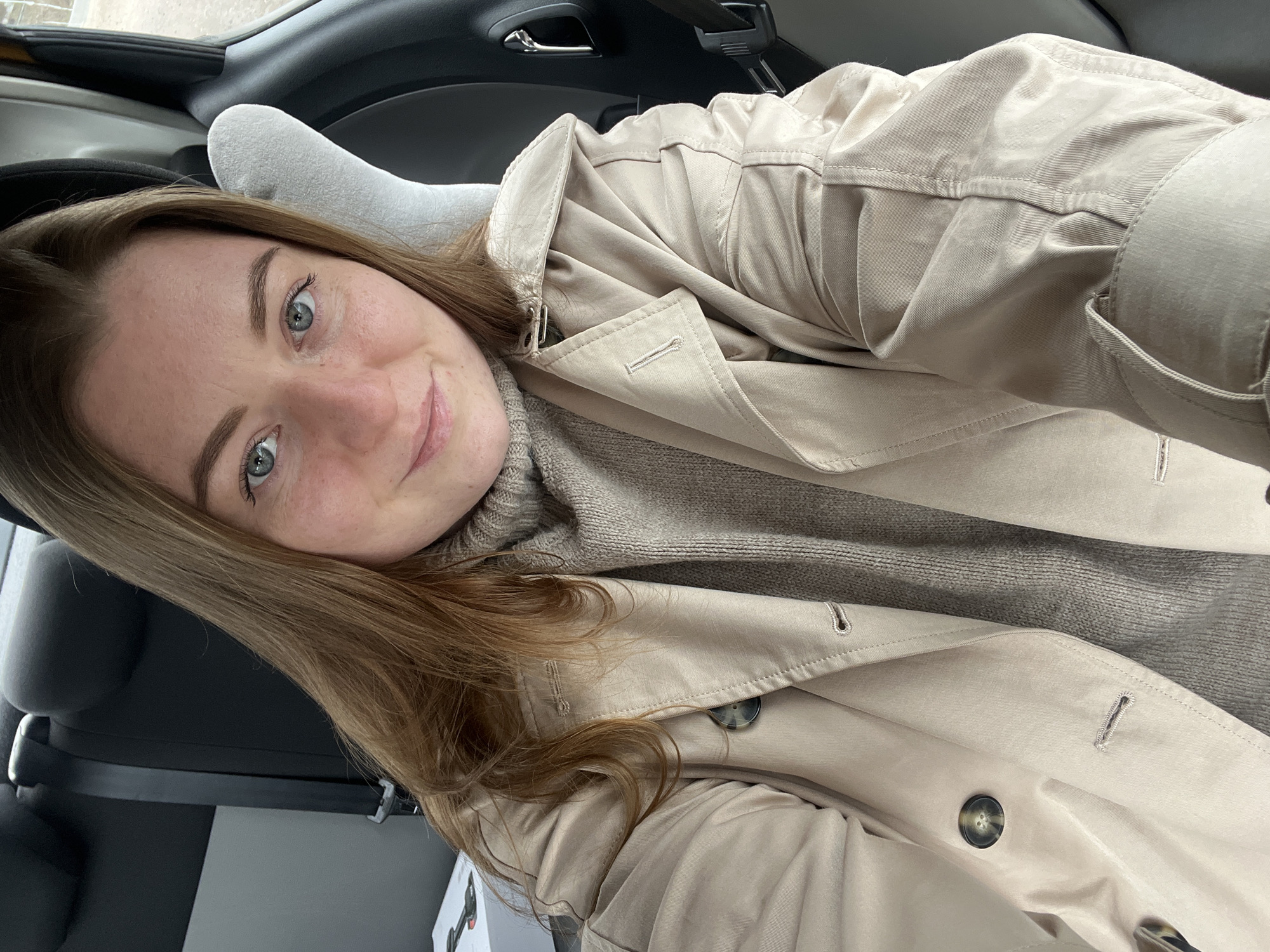 A woman is in a car, wearing a beige trench coat over a light brown turtleneck sweater. She has light brown hair and blue eyes. She is looking at the camera with a slight smile. A white sock is visible next to her head. The interior of the car is visible around her. The location is not determined.