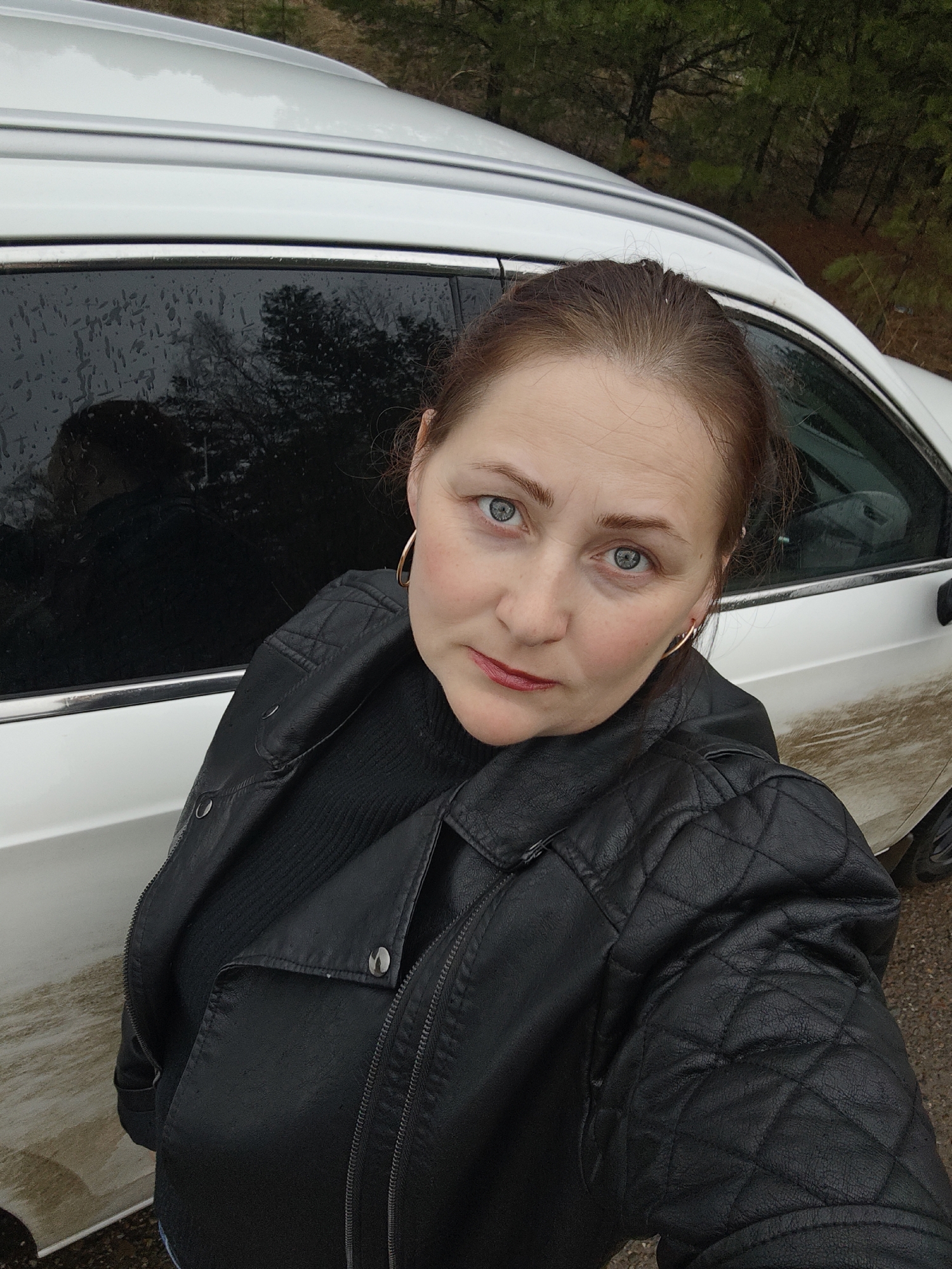 This is a selfie-style shot featuring a woman standing next to a white SUV in Chaikovsky, Russia. The woman, appearing to be in her 40s or 50s, has fair skin, light blue-grey eyes, and light brown hair pulled back. She is wearing a black quilted faux-leather jacket over a black ribbed sweater and small gold hoop earrings, with red lipstick. Her expression is neutral and direct as she looks into the camera.
The white SUV parked beside her shows visible signs of recent wet conditions; its side window is covered in raindrops, and the lower body panels are splattered with mud. In the background, beyond the car, are bare-branched trees, suggesting an outdoor, possibly wooded or rural setting. The lighting appears soft and diffused, indicating an overcast daytime. No visible text is present in the image.