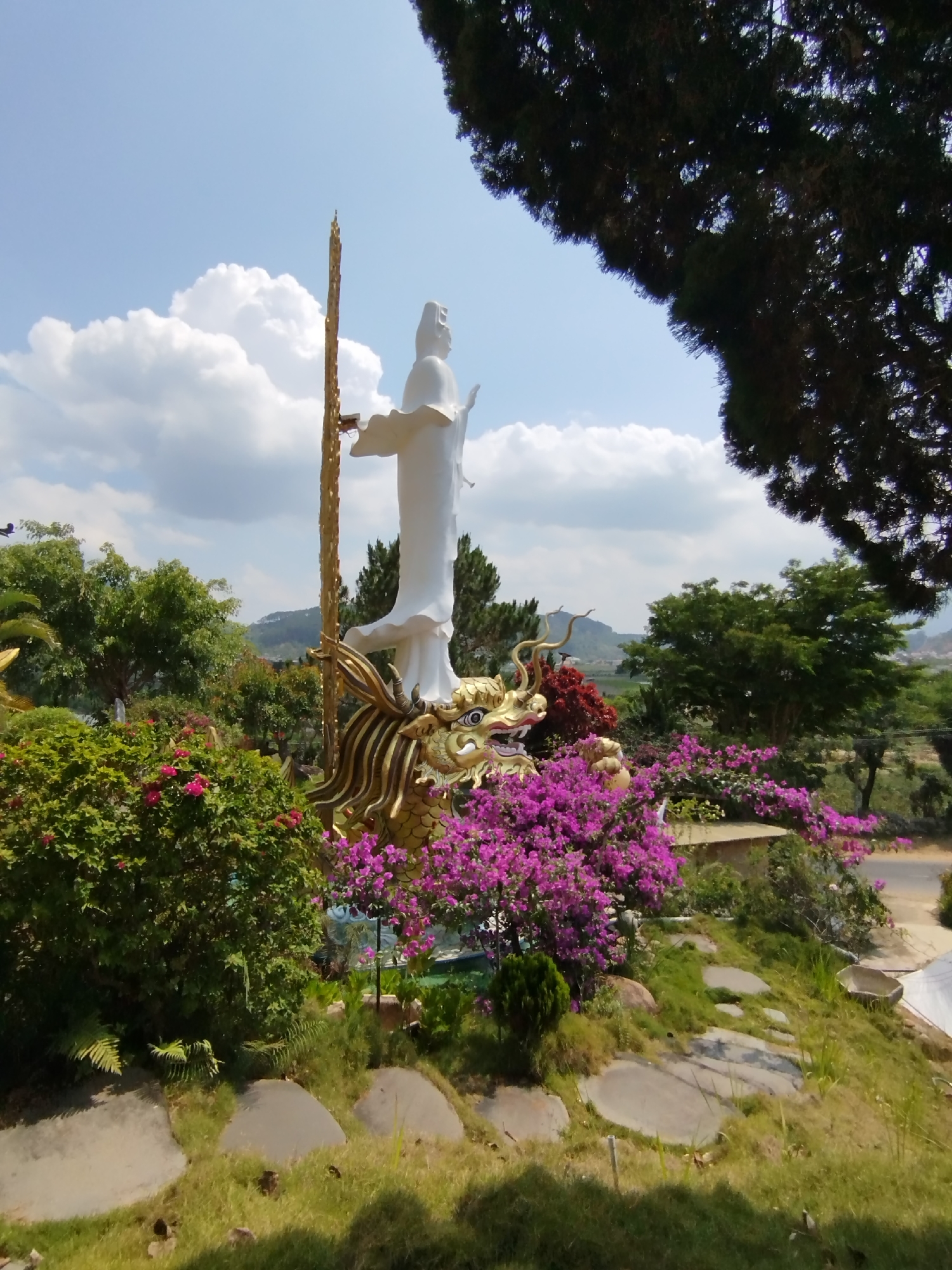 The image depicts a white statue of a deity, likely Guan Yin, standing atop a golden dragon statue in a garden setting. The statue and dragon are surrounded by lush greenery, including bushes with pink and red flowers. A golden staff stands beside the statue. The background includes trees, hills, and a partly cloudy sky. A portion of a tree branch is visible in the right foreground. Based on the prompt, the location is Diom A, Vietnam. The scene appears to be a religious or spiritual site.