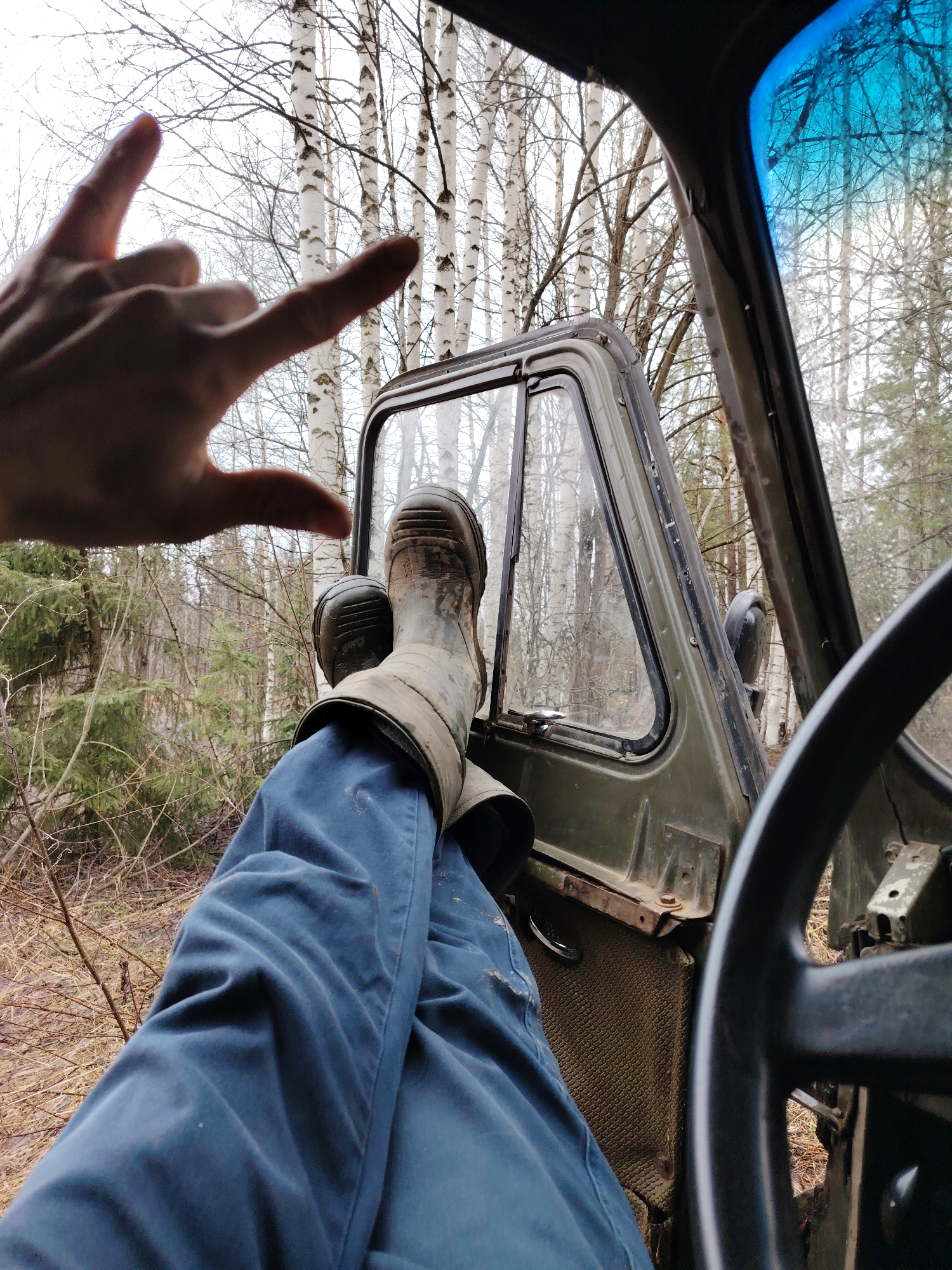 From within a vehicle, possibly in a wooded area near Markovskii, Russia, a person rests their legs, shod in dirty rubber boots, on the open door frame. The person wears blue pants. A hand is raised in a "rock on" gesture. Bare trees are visible through the open door and windshield. The steering wheel is partially visible in the foreground.