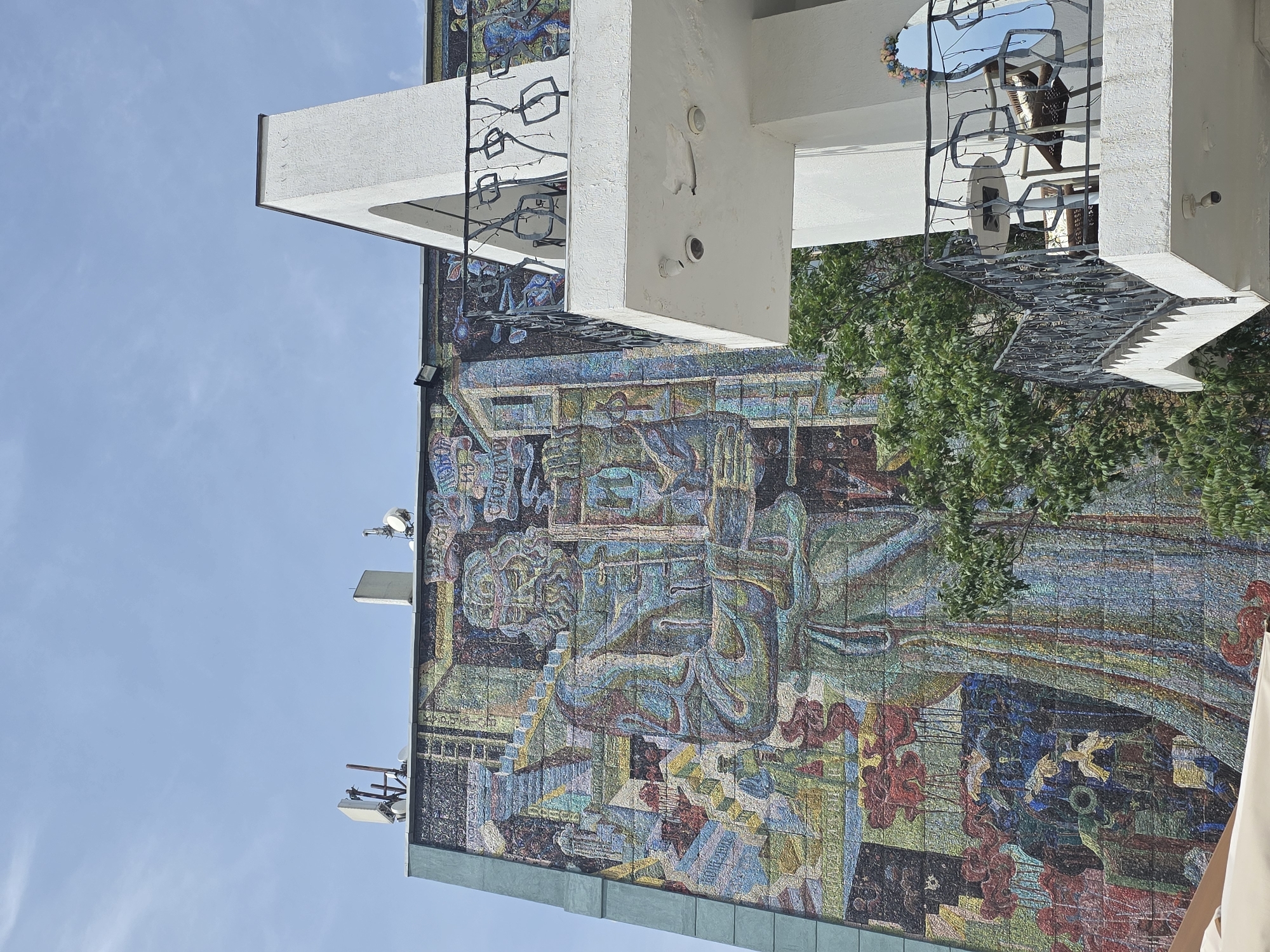 A building, possibly a high-rise, is partially visible in this daytime shot. The building's facade features a large mosaic mural in shades of blue, brown, green, and red. The mural depicts what appear to be human figures and other abstract shapes.
The building's architecture is modern, with white concrete protrusions and balconies. One balcony is decorated with metal latticework.
The sky is blue and clear, with the top left corner of the building framing the background. The perspective suggests the photo was taken from a low angle. A tree with green leaves partially obscures the right side of the mural. Antennas are visible on the building's edge.
This is the "Aurora" cinema in the city of Krasnodar, Russia.