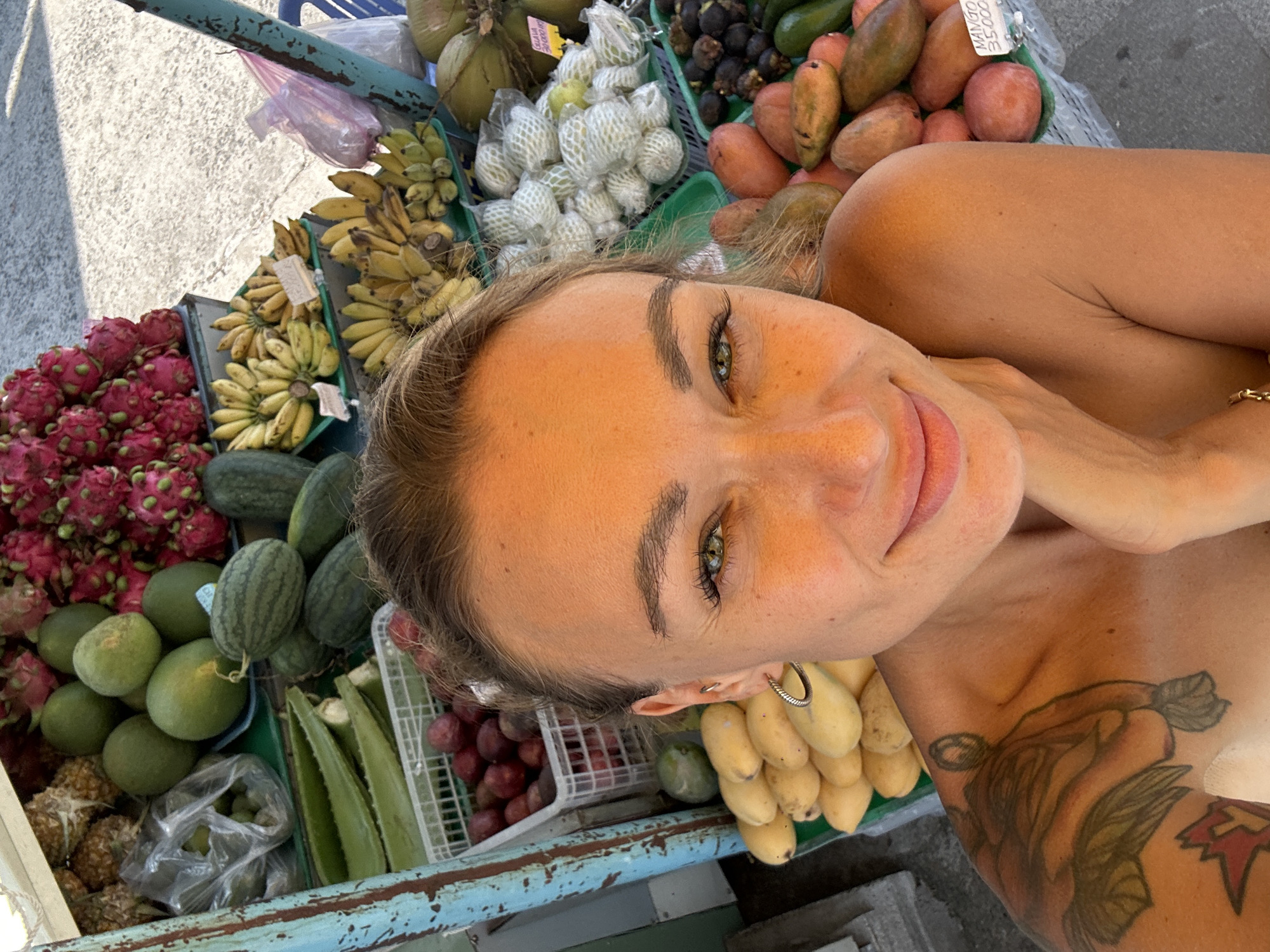 The image depicts a person with fair skin, brown hair, and green eyes posing next to a cart laden with various fruits, likely at a market. The fruits include dragon fruit, bananas, watermelons, mangos, coconuts, mangosteens, aloe vera, and grapes. The person has a tattoo on their shoulder and is wearing a silver hoop earring. The location is inferred to be in Phu Thuy Ward, Vietnam, based on the context provided.