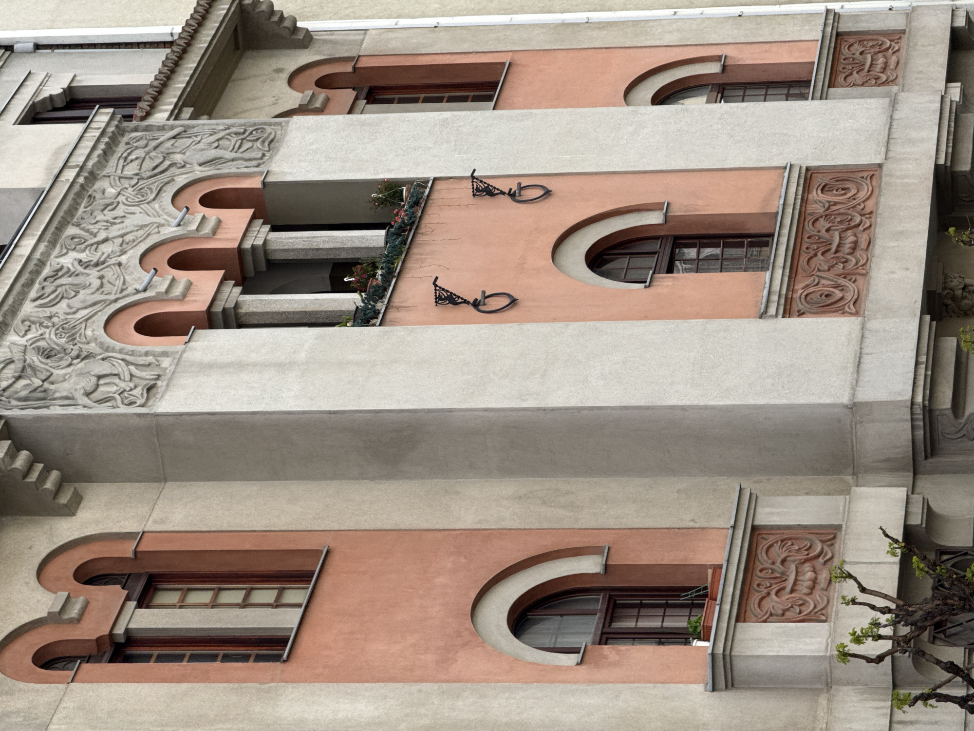 The image shows the façade of an Art Nouveau building in Belgrade, Serbia. The architecture features alternating horizontal bands of light grey concrete and reddish-pink stucco. The windows are rectangular and arched, with dark wood frames. Decorative elements include floral carvings on the concrete, iron balcony brackets, and a frieze above the balcony. Balcony has flowers.