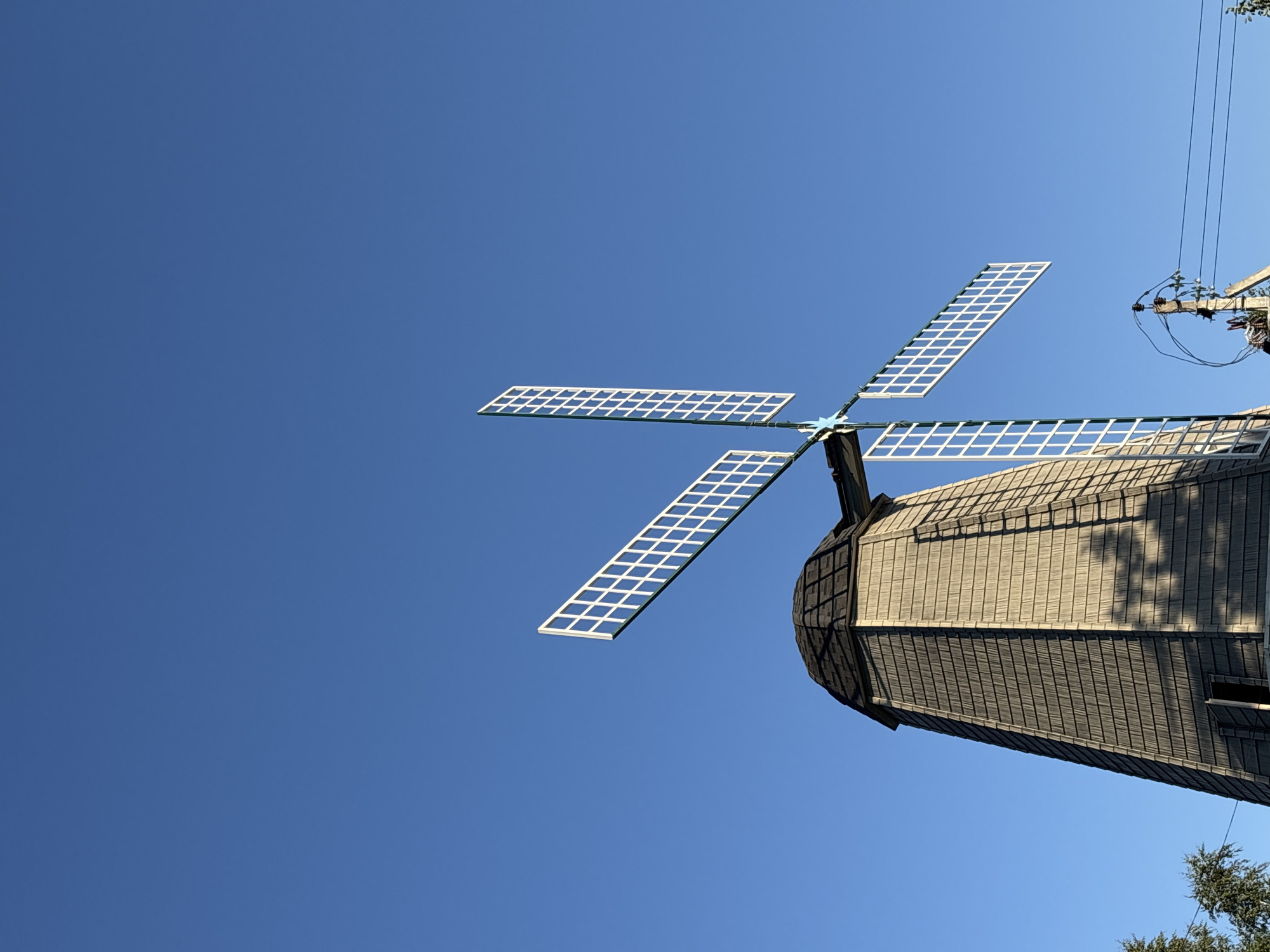 The image shows a Dutch-style windmill against a clear blue sky in the city of Romashkovo, Russia. The windmill has a conical, shingled tower and four white, lattice-like sails. One sail is prominently featured, angled towards the right of the frame, while another is visible pointing downwards on the left. The sails appear to be in a static position, not actively rotating. The lower portion of the windmill is visible, with some shadowed areas indicating texture from the shingles. In the lower right corner, a utility pole with wires is partially visible, suggesting the presence of infrastructure in the vicinity. The overall scene is tranquil, with the windmill being the central subject.