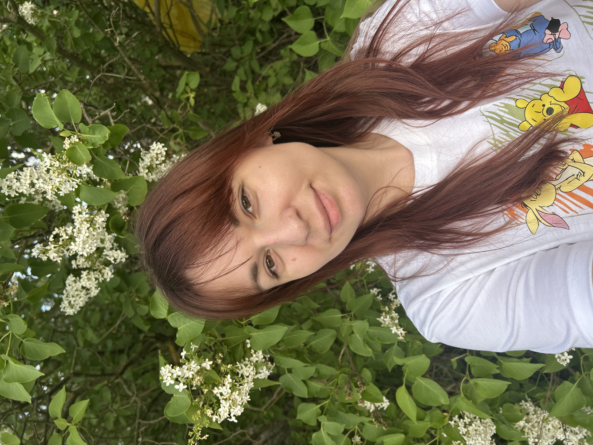 A woman with auburn hair stands near a flowering bush. She is wearing a white shirt with a cartoon character print. The bush has green leaves and small white flowers. The photograph was likely taken outdoors in a natural setting. There is no information in the image to determine that it was taken in the city of Фоки, Russia.
