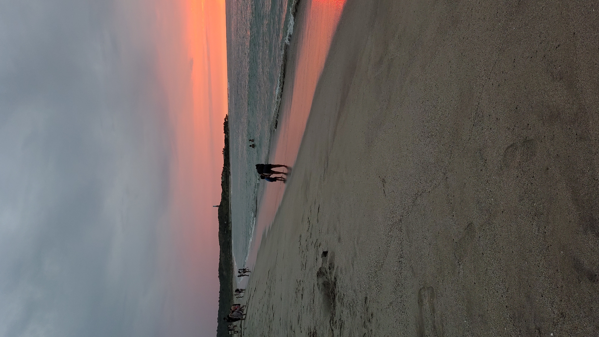 The image depicts a beach scene in Bocapan, Peru at sunset. The sky transitions from grey on the left to orange on the right. The ocean occupies the middle portion of the image. People can be seen walking on the beach, with silhouettes of a parent and child. The beach sand is a light brown color with footprints. Vegetation lines the horizon in the middle, and a structure resembling a lighthouse is visible.
