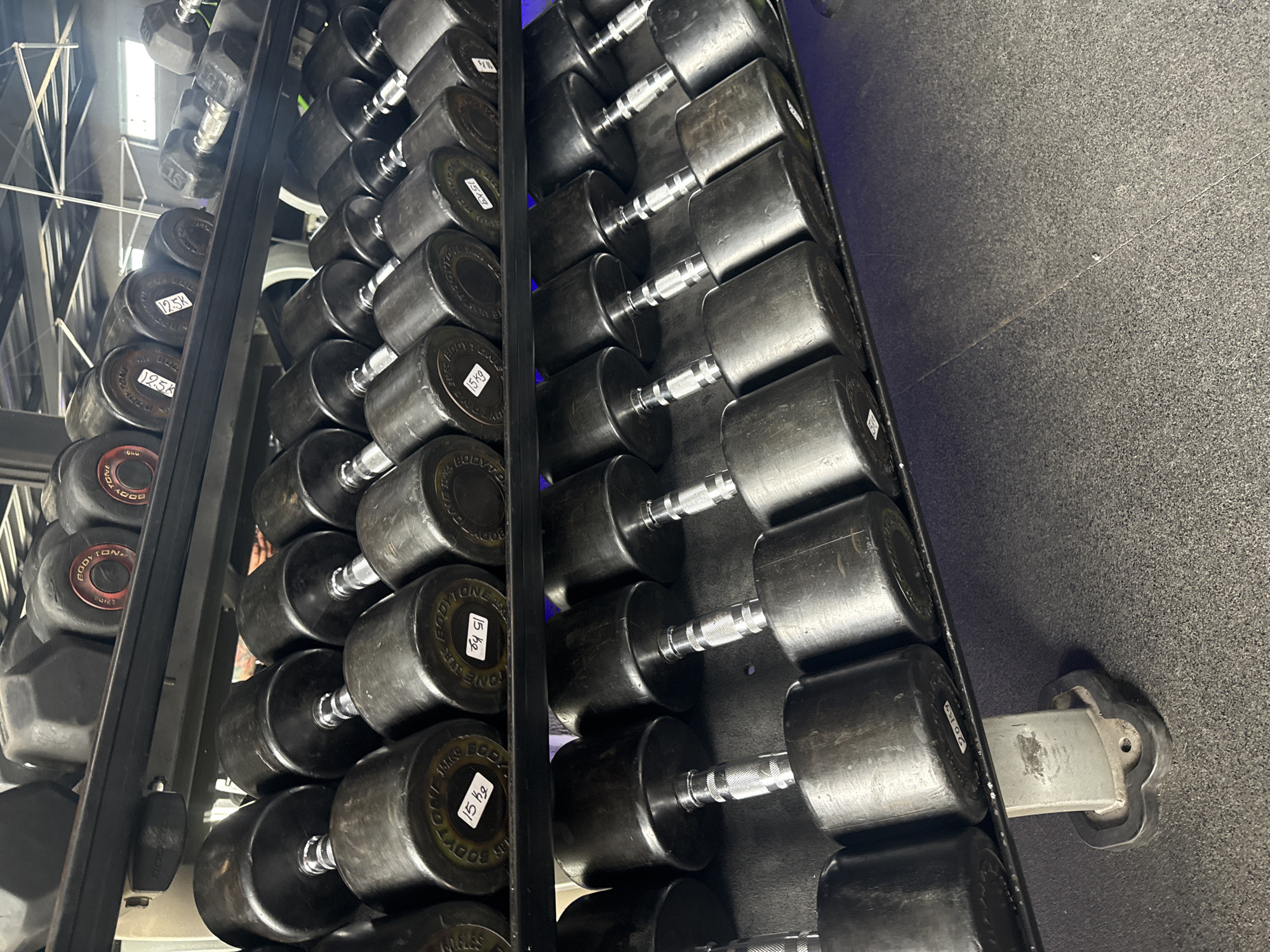 A rack of black dumbbells with silver handles is situated on a gray rubber floor, likely in a gymnasium or fitness center. The dumbbells range in weight, as indicated by the labels on the ends. Some labels read "15 kg", "12.5K" and "20lg" and "BODYTONE STAR MABLES" is seen on several of the dumbbells. The location is possibly in Puebla, Mexico. The rack is black and the background includes hints of the building's framework.