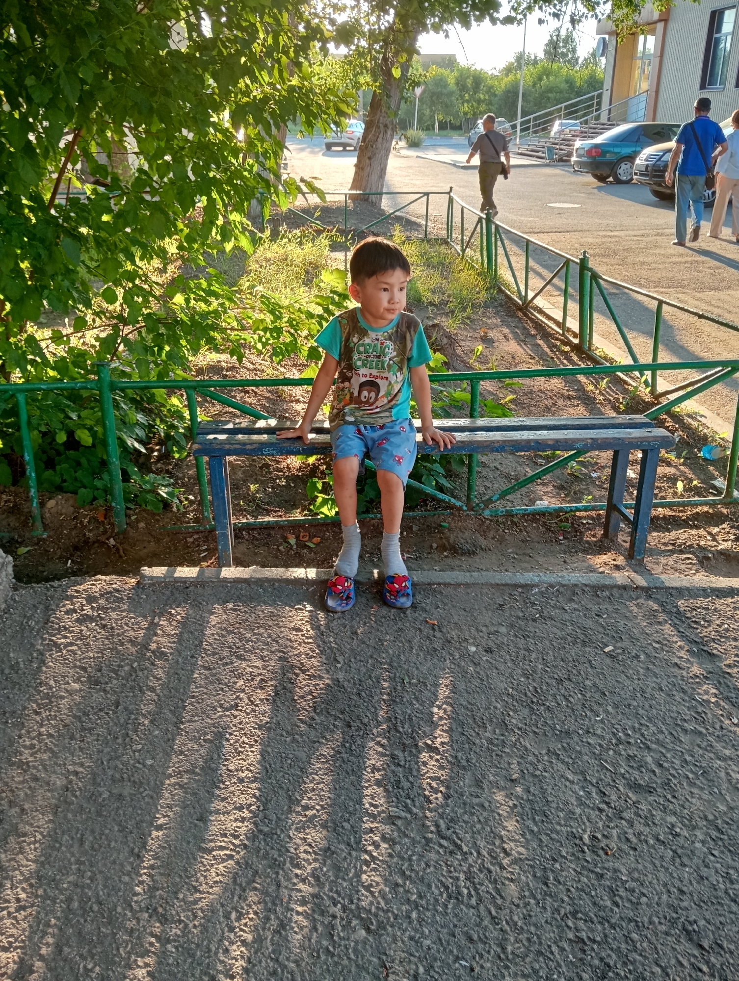 A young boy sits on a weathered blue wooden bench in the shade of a large tree, his legs dangling. He is wearing a teal t-shirt with a graphic print and blue shorts with a pattern, grey socks, and red slide sandals. A green metal fence runs behind him, separating the paved area from a patch of overgrown grass and dirt. In the background, several cars are parked on a paved surface, and other people are walking near a building. The lighting suggests it is late afternoon, casting long shadows from the trees and fence onto the ground.