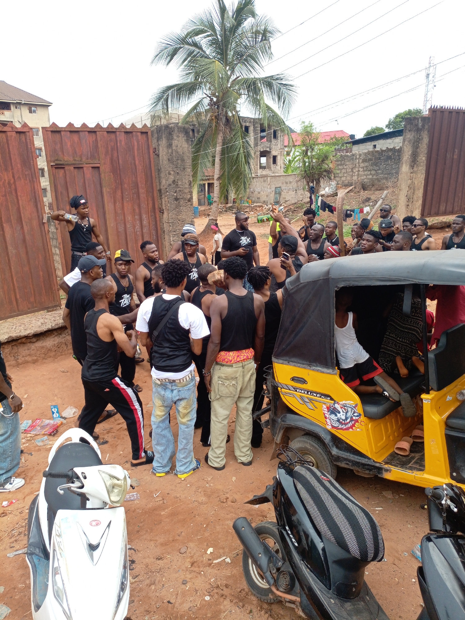 This media file captures a lively outdoor gathering of a large group of predominantly young men in an unpaved area in Onitsha, Nigeria. The ground is reddish-brown dirt, with various buildings in differing states of construction or repair visible in the background, along with a prominent tall palm tree and overhead power lines.
The central focus is a crowd of men, many dressed casually in black tank tops or vests, jeans, track pants, bandanas, and caps. Several of the black tank tops display the text "TRAP TALK." One man stands elevated atop a rusty brown metal gate, holding a clear plastic bottle, seemingly engaging or addressing the group below. The other men are clustered around, some looking at the man on the gate, others interacting amongst themselves.
Prominently featured in the foreground is a yellow tricycle, locally known as a "Keke," with "DELUXE PLUS" text and a wolf's head graphic on its side. A young man in a white tank top and red shorts is seated inside the Keke. Two scooters are parked nearby: a white one in the foreground with a partially visible red logo, and a black one (likely a "DIO" model, judging by the seat) further back. Discarded plastic bottles and wrappers are scattered on the ground.
The scene is brightly lit by natural daylight, indicating daytime and warm weather, suggested by the light clothing. Clothes are seen drying on a makeshift line in the distant background. The atmosphere appears social and dynamic.