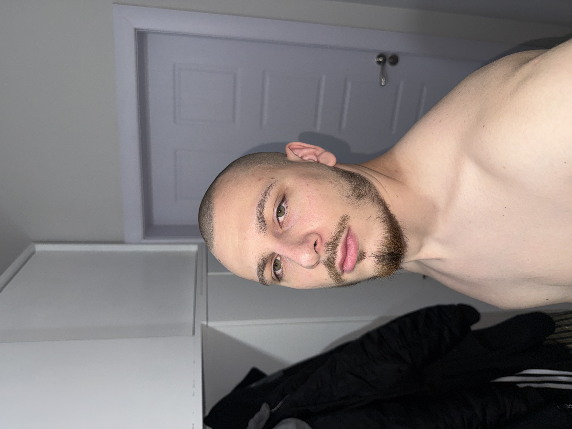 A person with a shaved head and a beard is shown in a close-up shot. The person has light skin and is bare-chested. The background consists of a white door and a white cabinet. Black clothing is visible at the bottom of the frame. There is no information to suggest that the photograph was taken in Stara Zagora, Bulgaria.