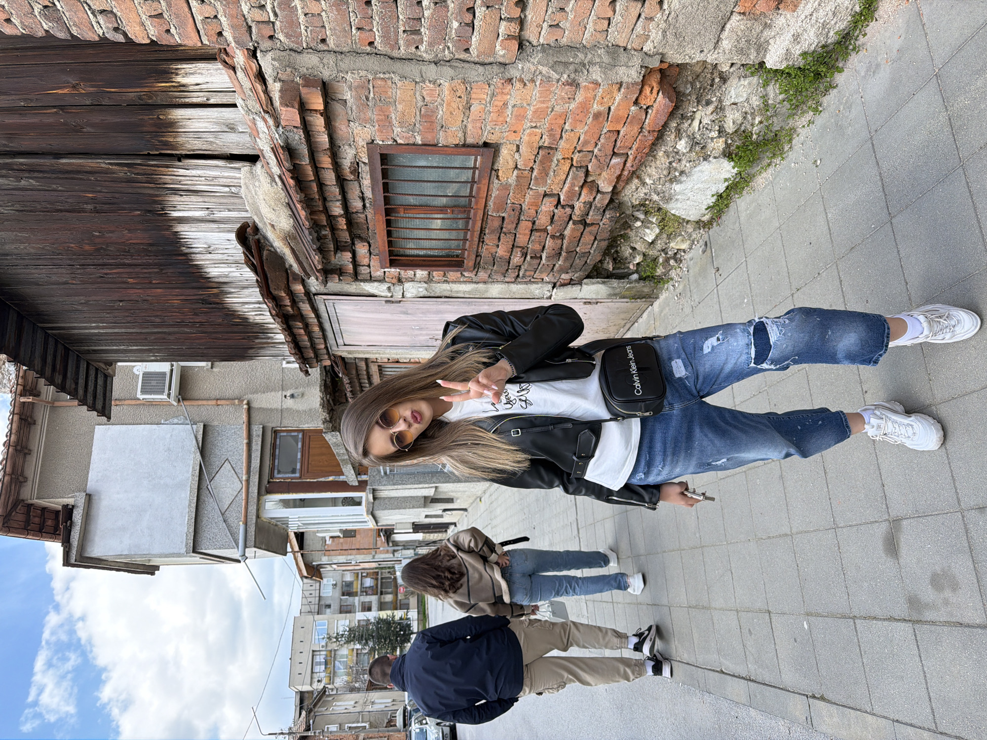 The image depicts a street scene in Panagyurishte, Bulgaria. A young woman wearing sunglasses, a black leather jacket, a white t-shirt, and ripped blue jeans poses for a photograph, holding up a peace sign. She has a black Calvin Klein jeans shoulder bag. Two other individuals are walking behind her on the sidewalk: one wearing a brown jacket and jeans, and another wearing a dark blue jacket and khaki pants. The setting is an urban environment with buildings and streets visible in the background. The architecture is a mix of old brick and modern structures. There is a cloudy sky visible above the buildings. The woman is standing on a paved sidewalk next to an old brick building.