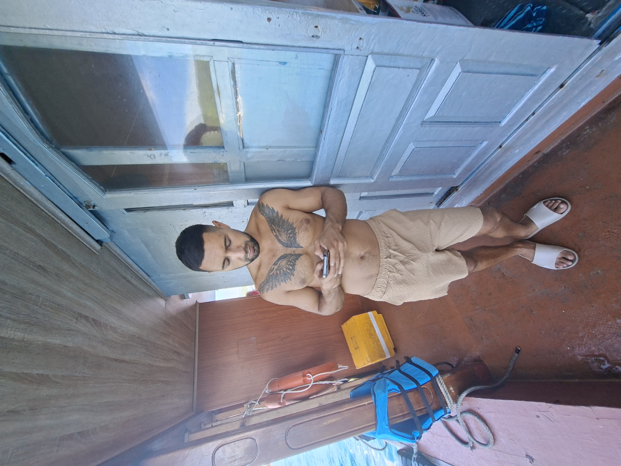A shirtless man with a wing tattoo on his chest stands on the deck of a boat, looking at his phone. He is wearing beige shorts and white flip-flops. The boat has a wooden interior and a light blue door with glass panels. Orange life preservers and a blue life jacket are visible on the left side of the boat. The boat appears to be docked or anchored, with a body of water visible on the left. The setting is consistent with a location in the city of Nam Nha Trang Ward, Vietnam, which is known for its coastal activities and boat tours.