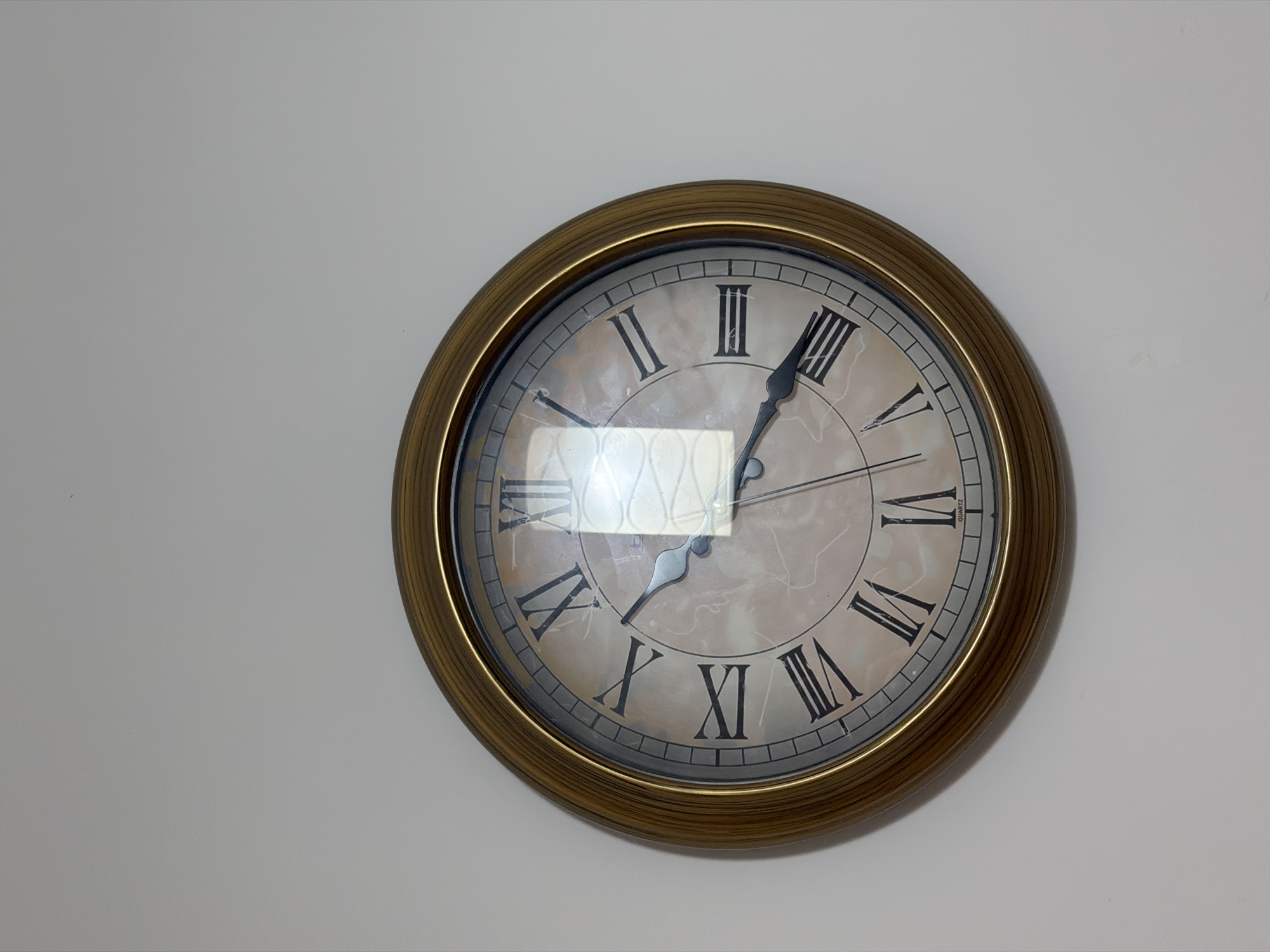 A circular, analog clock with a gold-colored frame hangs on a plain white wall. The clock face features Roman numerals and has a light, marbled pattern. The hour hand is pointing approximately to the 5, and the minute hand is pointing approximately to the 7. There is a reflection on the glass of the clock, showing a diamond pattern. The clock appears to be mounted on a wall indoors, with no visible indication of its specific location within Budva, Montenegro.