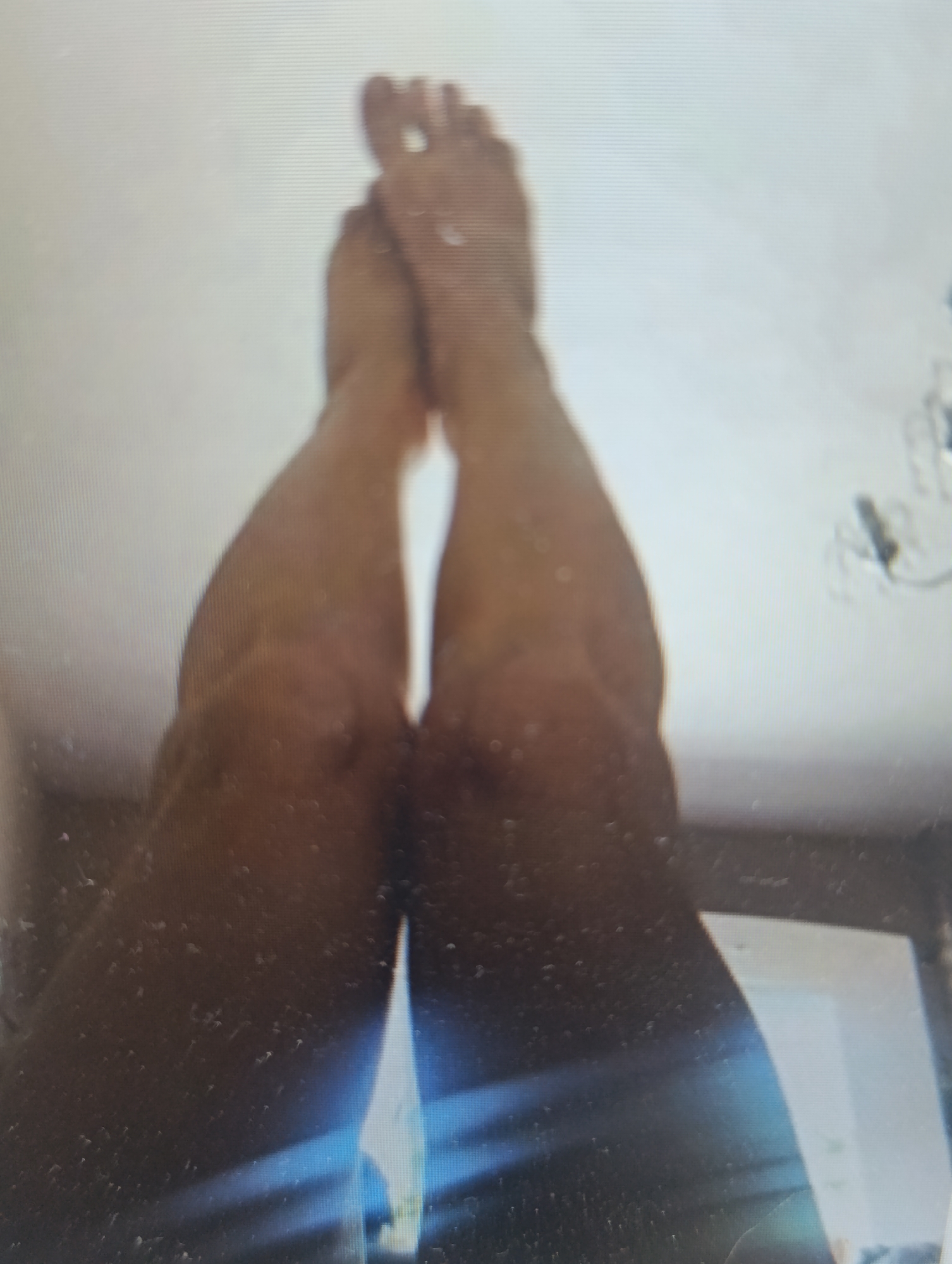 The image shows a close-up view of two human legs from the knees down, with the feet visible at the top. The legs are positioned in a V-shape, with the knees bent and the feet facing upwards. The skin tone appears to be light brown. The lighting is bright, suggesting an indoor setting with overhead illumination. The background is largely white, with some indistinct shapes and light sources visible in the upper right and bottom right corners. There is some visual noise and blurriness throughout the image, possibly due to low resolution or a moving subject. No specific activity or event is clearly depicted, beyond the pose of the legs. There is no visible text.