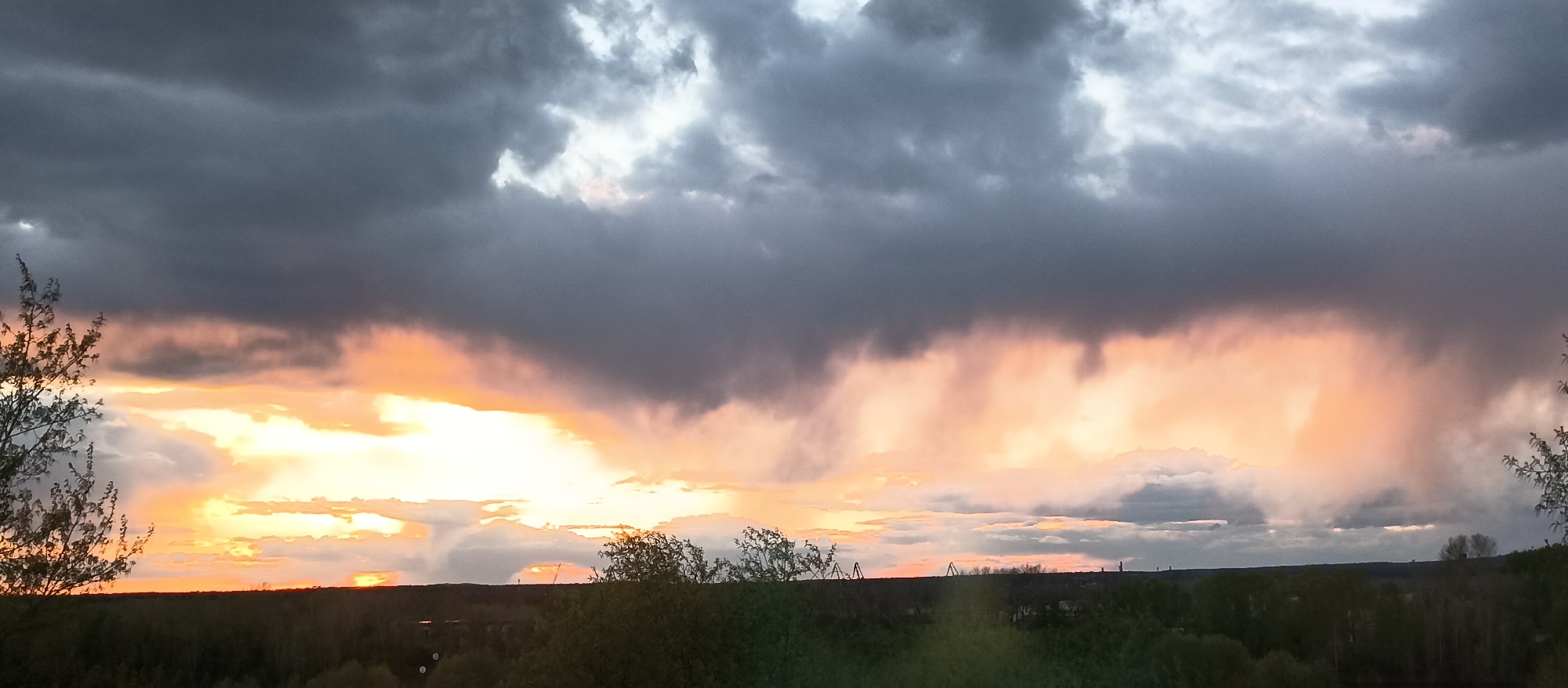 The image captures a landscape under a dramatic sky. The sky is dominated by dark, heavy clouds, some tinged with shades of gray and white above, while others closer to the horizon have hints of orange and yellow from the setting sun. Patches of light peek through the clouds.
Below the sky, a landscape with trees and vegetation is visible, forming the lower portion of the image. The trees appear as silhouettes.
The photograph seems to be taken from a high vantage point. There are no people visible in the image.