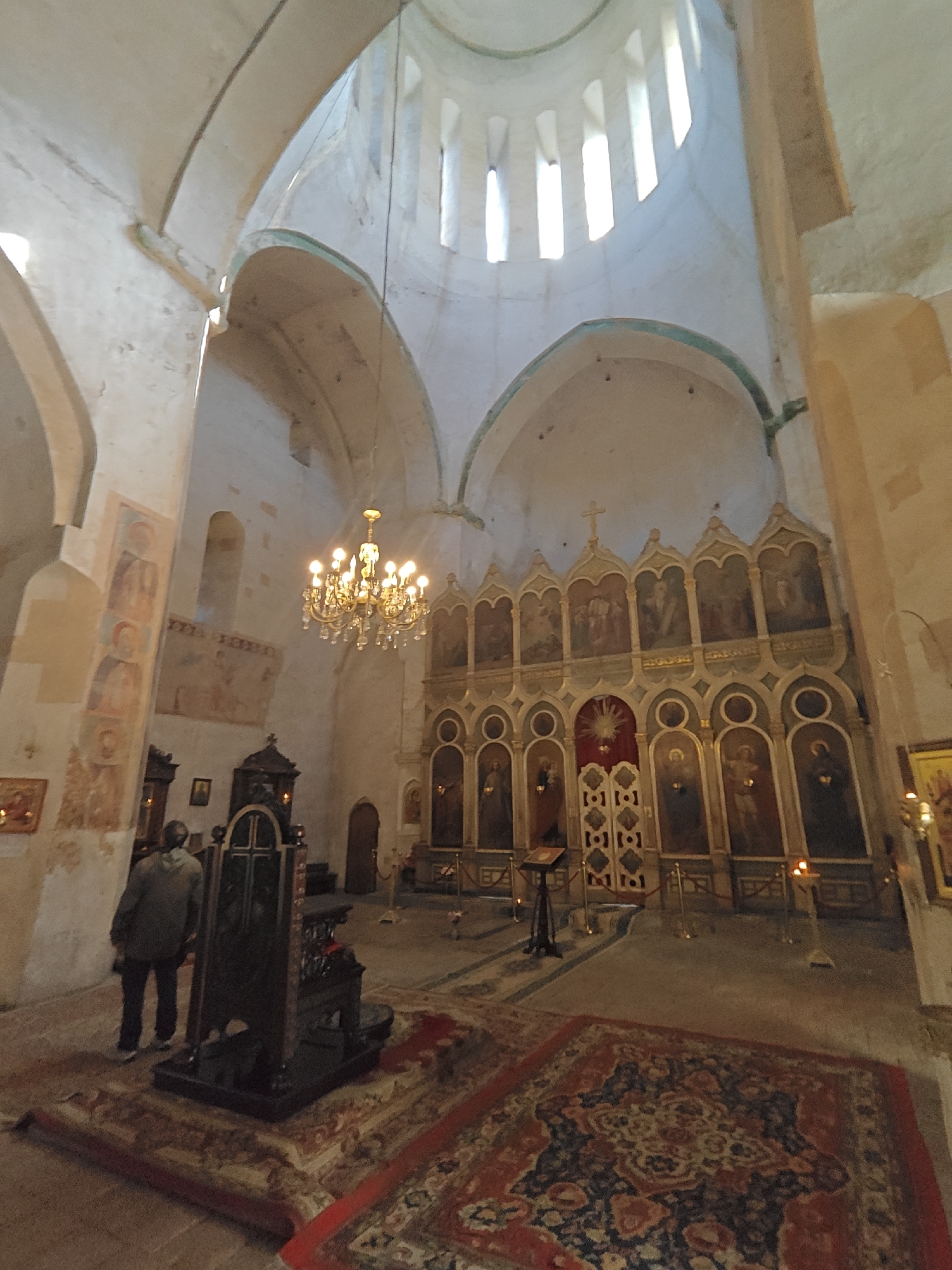 This image offers an interior view of a historic church or monastery in Pkhundavi, Georgia, characterized by its high vaulted ceilings, arches, and a central dome. The walls are painted in a light cream or beige, with some arches featuring a faded greenish-blue trim.
At the front of the sanctuary, a prominent, multi-tiered iconostasis stands, richly adorned with religious icons and paintings, featuring gold and blue decorative accents, and a central double door. A large, ornate golden chandelier with numerous lights hangs from the center of the dome, providing artificial illumination. The dome itself is pierced by several tall, narrow windows, allowing natural daylight to stream into the space.
Faded frescoes or murals depicting religious figures are visible on the high walls, particularly on the left side, indicating the building's ancient heritage. The floor is covered with large, intricately patterned carpets, predominantly red and blue with floral and geometric designs.
In the left foreground, a single person is seen from behind, seemingly observing the surroundings or engaged in devotion. Near them, a dark, carved wooden structure, possibly a reliquary stand or seat, is placed on the carpet. Several small stands with lit candles are scattered near the iconostasis, contributing to the serene and spiritual atmosphere. The scene captures a quiet moment within this sacred and historical setting, likely during daylight hours due to the natural light. No legible text is visible.