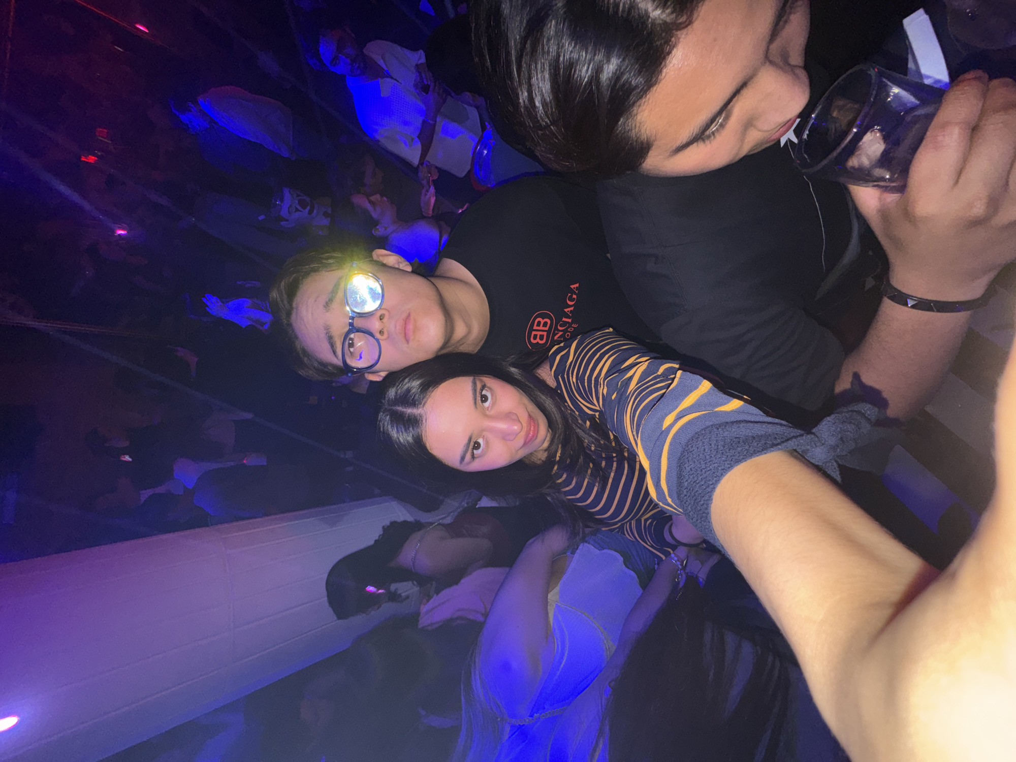 Three people are the primary subjects of this vertically-oriented, high-angle shot taken in what appears to be a nightclub environment.
The person closest to the camera is a young woman with dark hair, wearing a navy blue and yellow striped top. Her gaze is directed towards the camera.
To her left is a young man wearing glasses and a black t-shirt with a "BB" logo. He is looking slightly upwards.
To the right of the woman and above the man, a second man is holding a glass to his lips, as if drinking from it. He has short, dark hair, wears a black t-shirt, and has a bracelet on his left wrist.
The background is dark and blurry, with indistinct shapes of people. There are patches of blue and purple lighting. A white object stretches across the bottom left of the image. The exact location in San Bernardino Tlaxcalancingo, Mexico is unidentifiable from the image.