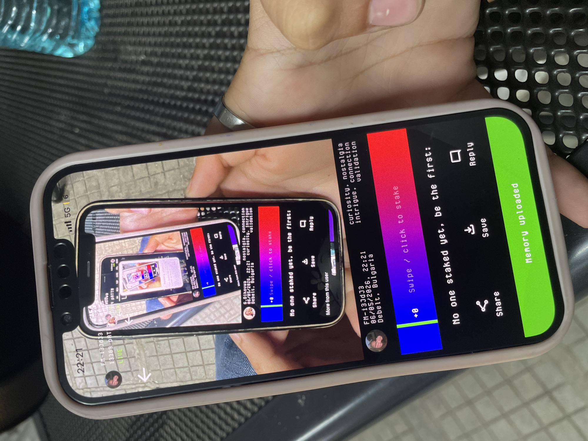A person's right hand, with a silver ring on the ring finger, holds a light-beige cased smartphone. The phone's screen displays a dark-themed application, which prominently features a recursive image of a smartphone screen within itself. The outermost phone's status bar indicates "22:21" and "5G" connectivity.
The primary application interface shows a profile labeled "FM-i3JdJ3" with a small profile picture, dated "06/05/2026, 22:21" and located in "Debelt, Bulgaria." Associated text lists "curiosity, nostalgia intrigue, connection validation." A horizontal blue-to-red gradient bar includes the text "+0 Swipe / click to stake." Below this, "No one staked yet, be the first:" is displayed, followed by icons for "Share," "Save," and "Reply." At the bottom of the screen, a prominent green button states "Memory uploaded."
Within this primary application, the recursive image shows another phone screen, displaying an almost identical application interface. This inner interface attributes content to "G.Dikouwa" with the same date, time, and location (Debelt, Bulgaria), along with similar descriptive tags and staking options.
In the background, a blue plastic bottle is partially visible on the upper right, and the surface below the phone is dark and textured, possibly a woven fabric or perforated material.