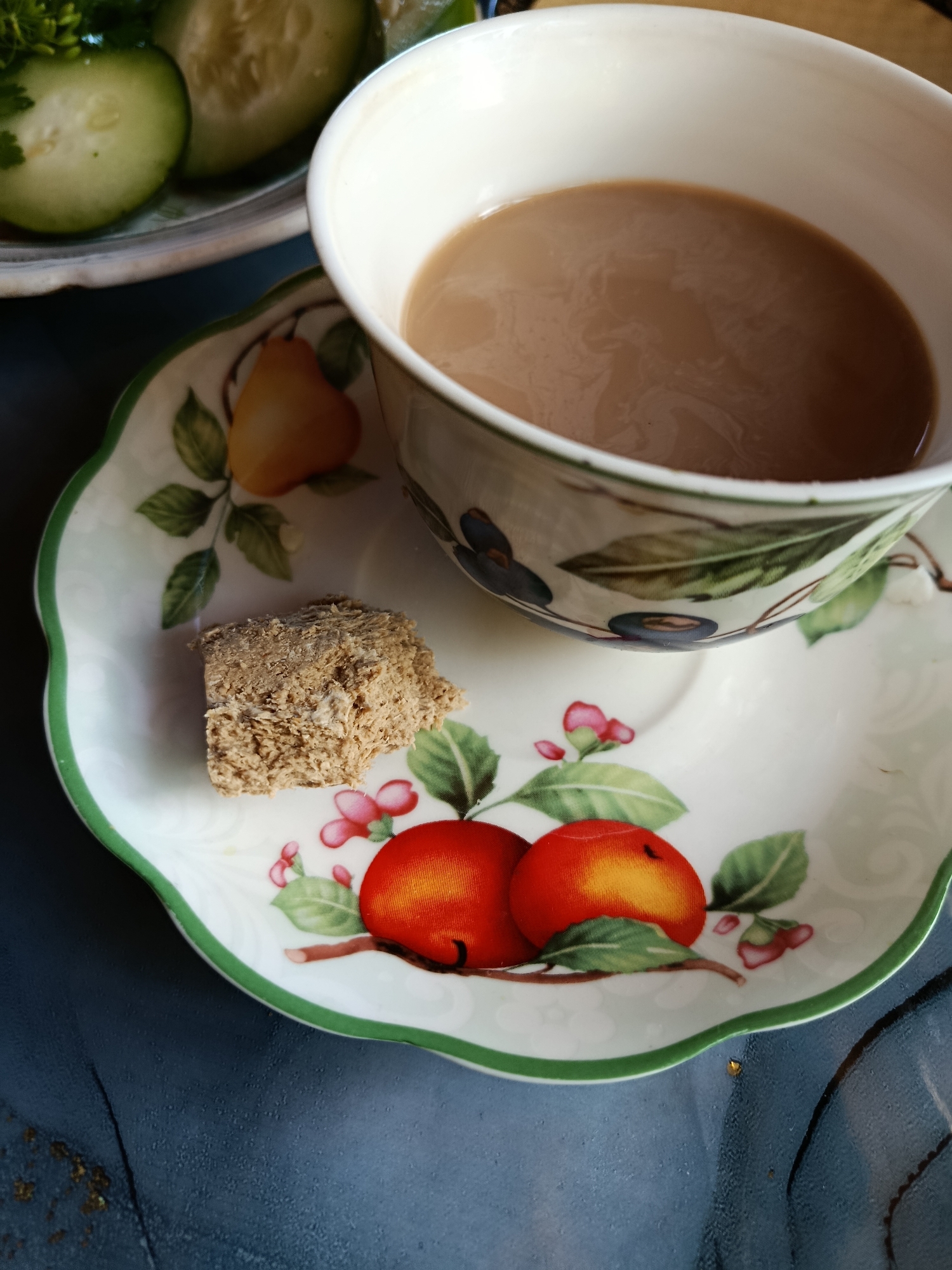 A close-up, overhead shot shows a cup of milky tea on a decorative saucer featuring red peaches and green leaves. A small piece of food, possibly halva, rests on the saucer near the tea cup. In the upper left corner, sliced cucumbers are visible on a plate. The saucer and cup are white with a green rim, and the saucer's design is vibrant and detailed. The background is a dark, textured surface, possibly a table. The lighting is natural, casting soft shadows. The scene appears to be set indoors.