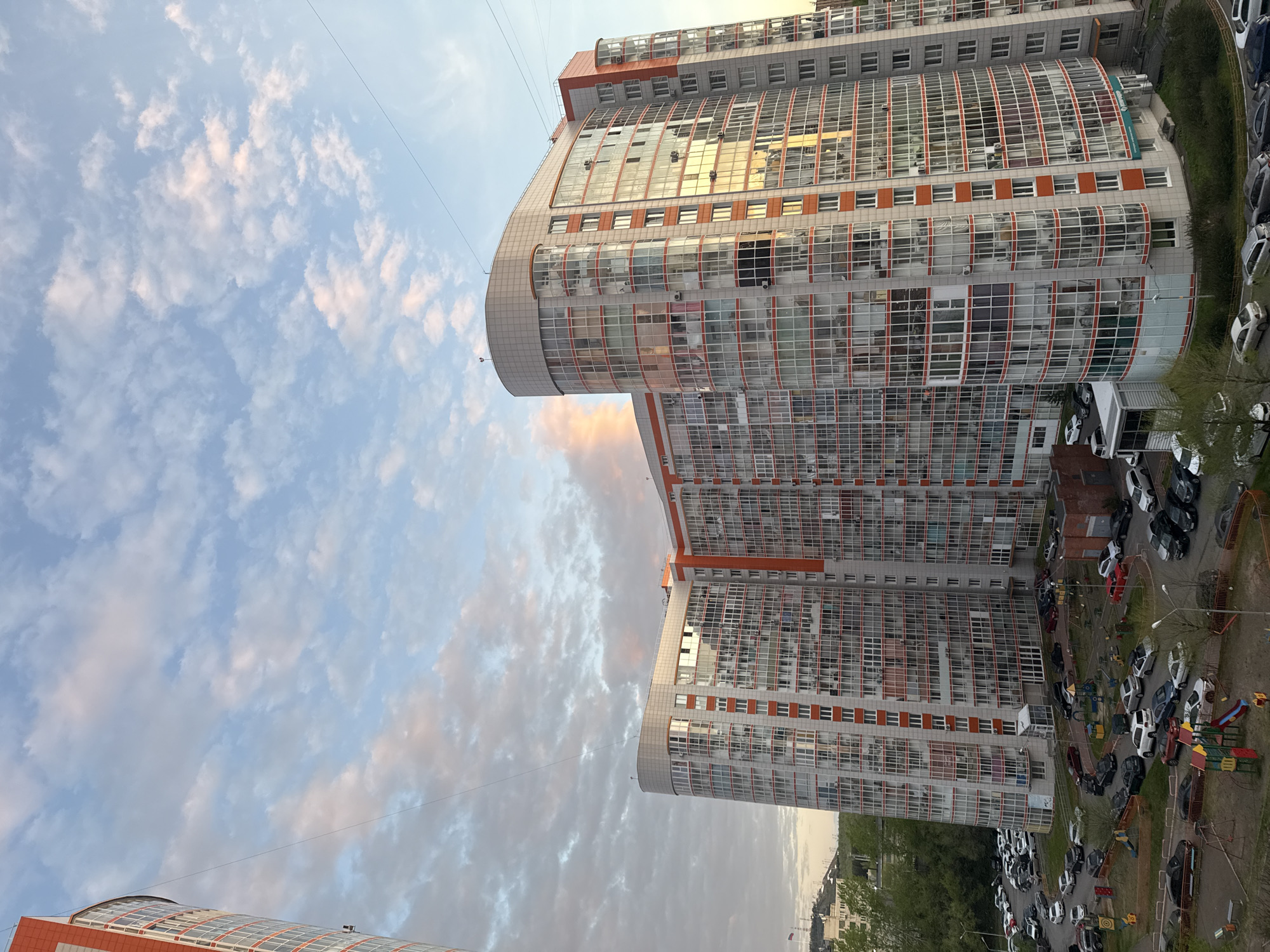 The image shows a tall, modern building complex in Krasnoyarsk, Russia, with many windows. The building has an orange and gray color scheme. A large parking lot filled with cars is adjacent to the building. The sky is partially cloudy, with some pink hues indicating either sunrise or sunset. Power lines are visible.