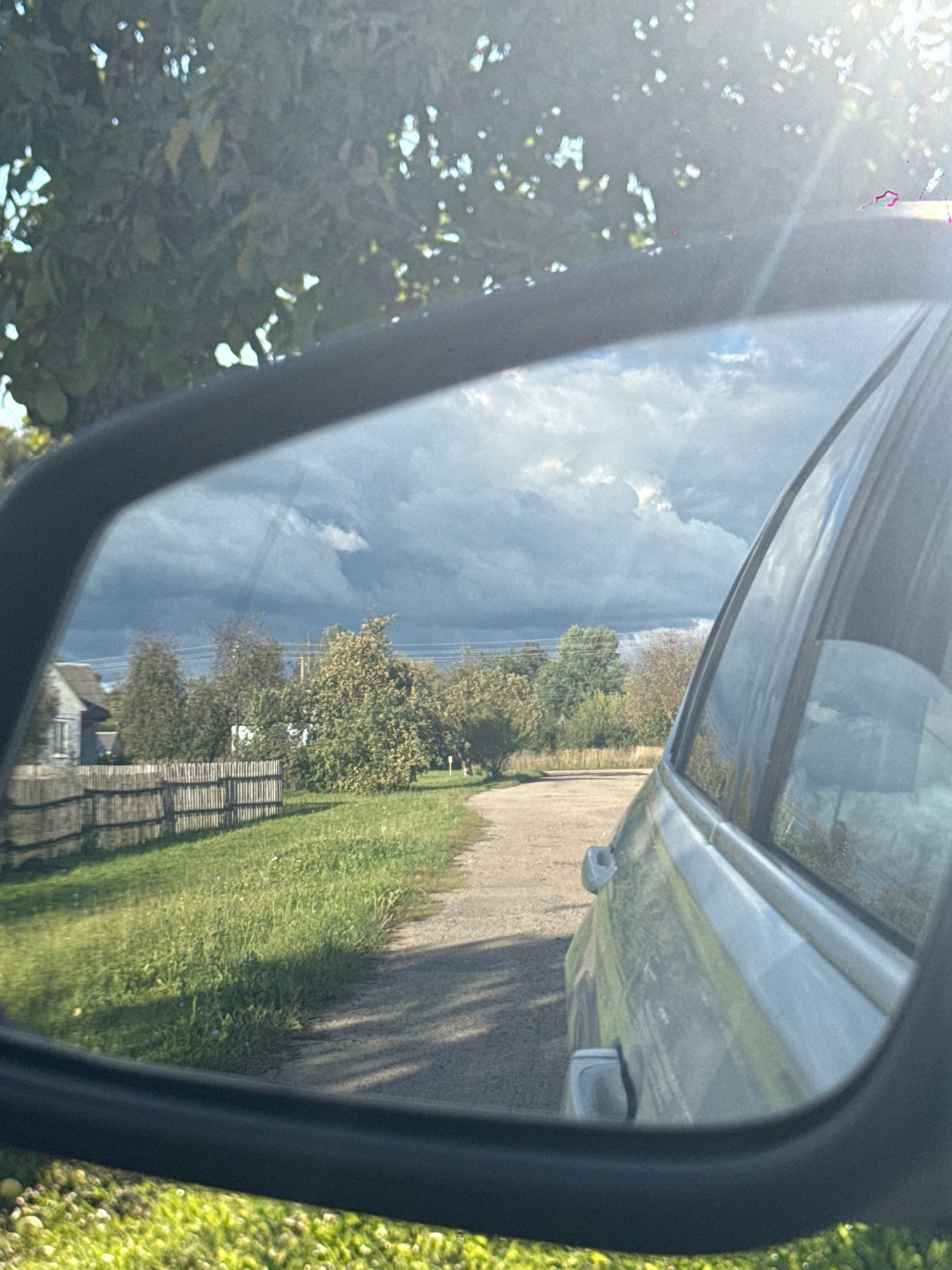 The image is a reflection in a car's side mirror, taken from within the car. The reflection shows a rural scene with a dirt road curving to the left, flanked by a grassy area on the left and a gravelly shoulder on the right. In the distance, there are trees and a wooden fence. A small, light-colored building with a dark roof is visible behind the fence. The sky is filled with dark, dramatic clouds. The foreground of the reflection includes some greenery. The car's exterior is visible in the right side of the frame, appearing to be a light metallic color. The location is identified as Slawharad, Belarus.