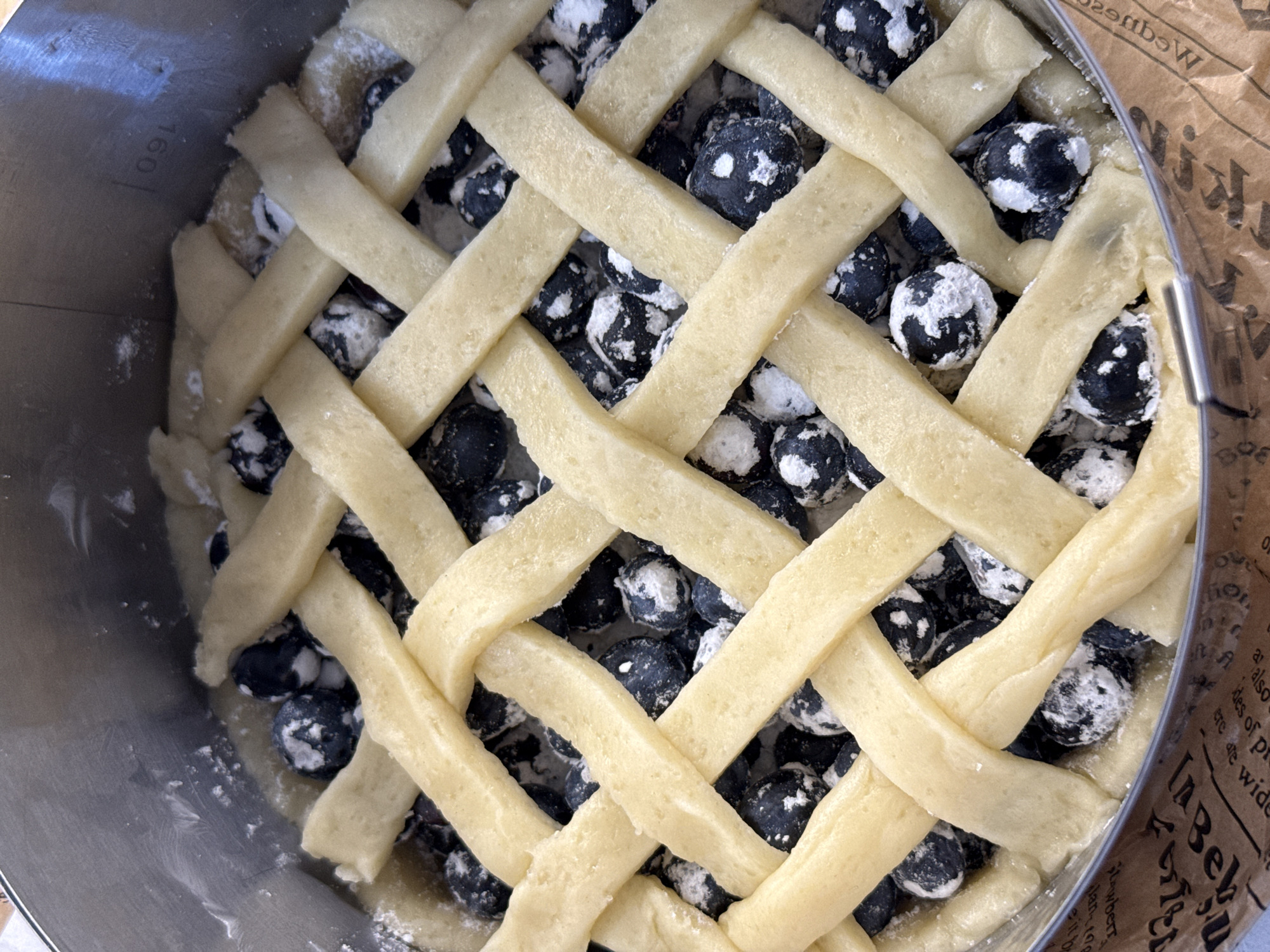 A close-up, overhead shot captures a blueberry pie being prepared in a round, stainless steel pie tin. The pie crust is elaborately latticed with pale yellow dough strips, woven over a generous filling of dark blue blueberries. Some of the blueberries appear dusted with a white powder, possibly flour or sugar. The pie tin itself is clean, with some white powder visible on its rim. The pie is sitting on a brown paper, which has printed text on it. There are no people visible in the image, and no specific geographic indicators that would place it in Lyubertsy, Russia.