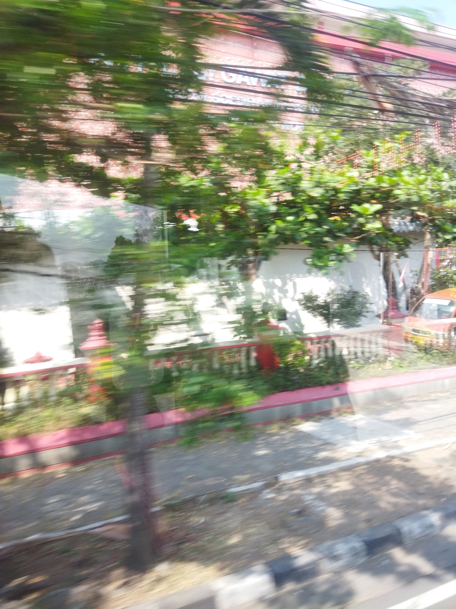 The image is a blurred, horizontal, medium shot capturing a view from a moving vehicle, likely a car or bus, through a window. The scene depicts a street in the city of Wonogiri, Indonesia, with a focus on vegetation, a fence, and a building in the background. The motion blur suggests rapid movement.
In the foreground, a gray and black paved curb is visible, with a sidewalk adjacent to it. The sidewalk is composed of compacted dirt and gravel, with scattered leaves and shadows.
Beyond the sidewalk is a pink and gray fence, with white vertical posts topped with decorative red elements. Lush green foliage, including trees and bushes, is visible on the other side of the fence. Some of the plants have small reddish-pink flowers.
In the middle ground, a white wall forms the backdrop behind the fence and vegetation. The wall appears to be part of a building. Shadows from the trees are cast upon it. A red and orange-colored car is parked on the right side of the image, partially obscured by foliage and motion blur. A flag, possibly the Indonesian flag, is visible on a pole behind the car.
The background shows a building with a red roof and a pink facade, covered in what appears to be a pattern of small red and white elements. A dense network of electrical wires crisscrosses the upper portion of the image, adding to the sense of urban environment. The sky is a pale blue.