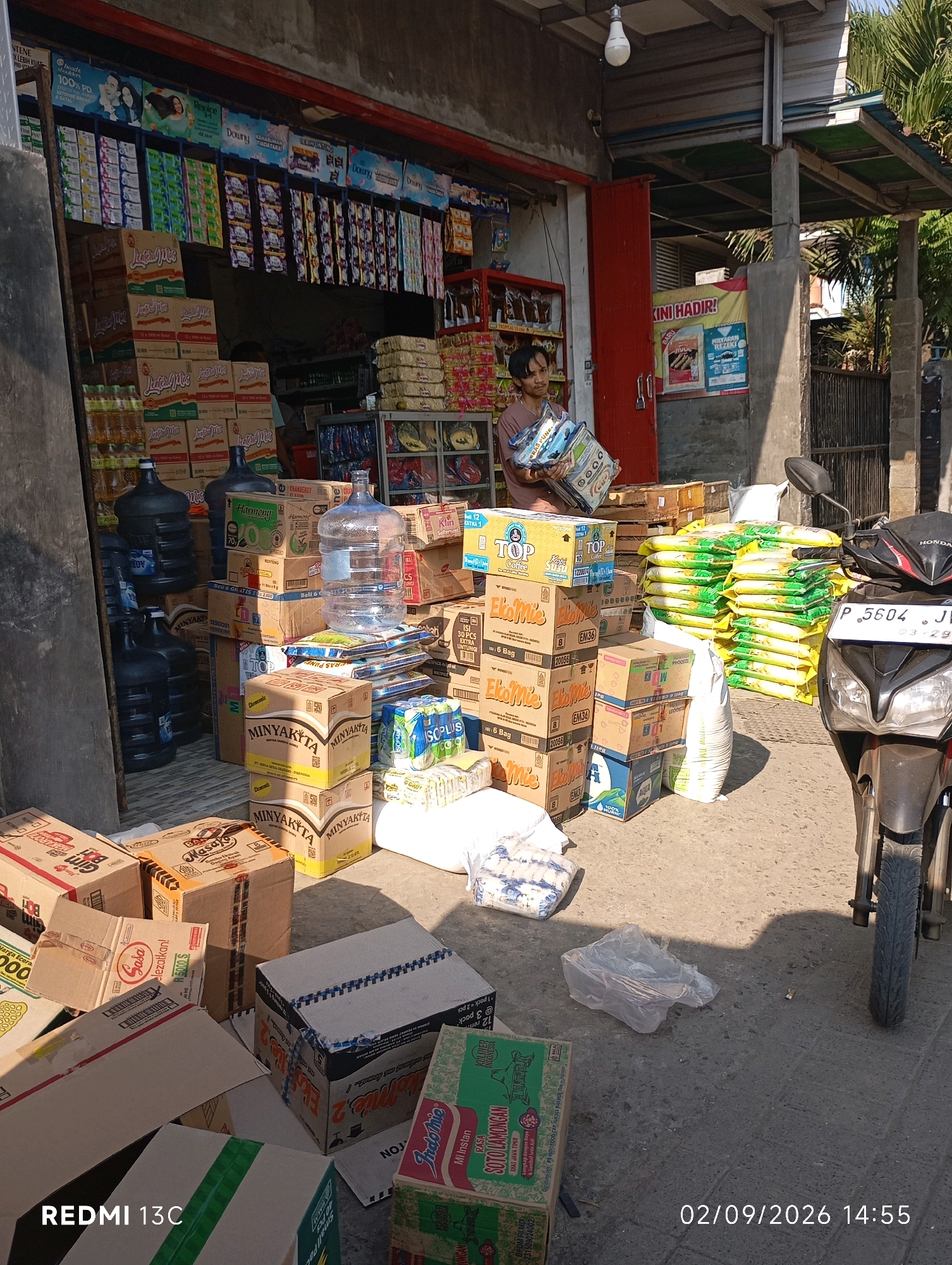 The image depicts a street-level view of a small store in Rambipuji, Indonesia, on a sunny day. The store appears to be a general goods or minimarket. In the foreground, there are various boxes of merchandise, including bottled water, instant noodles, and cooking oil. A motorcycle is parked on the right side of the frame. A person is visible inside the store, partially obscured by boxes, carrying several items. The store's exterior features shelves stocked with products, and banners advertising various brands are displayed. The environment is a typical Indonesian street scene with concrete flooring and a building with a corrugated metal roof. The date and time stamp on the image indicates September 2, 2026, at 14:55.