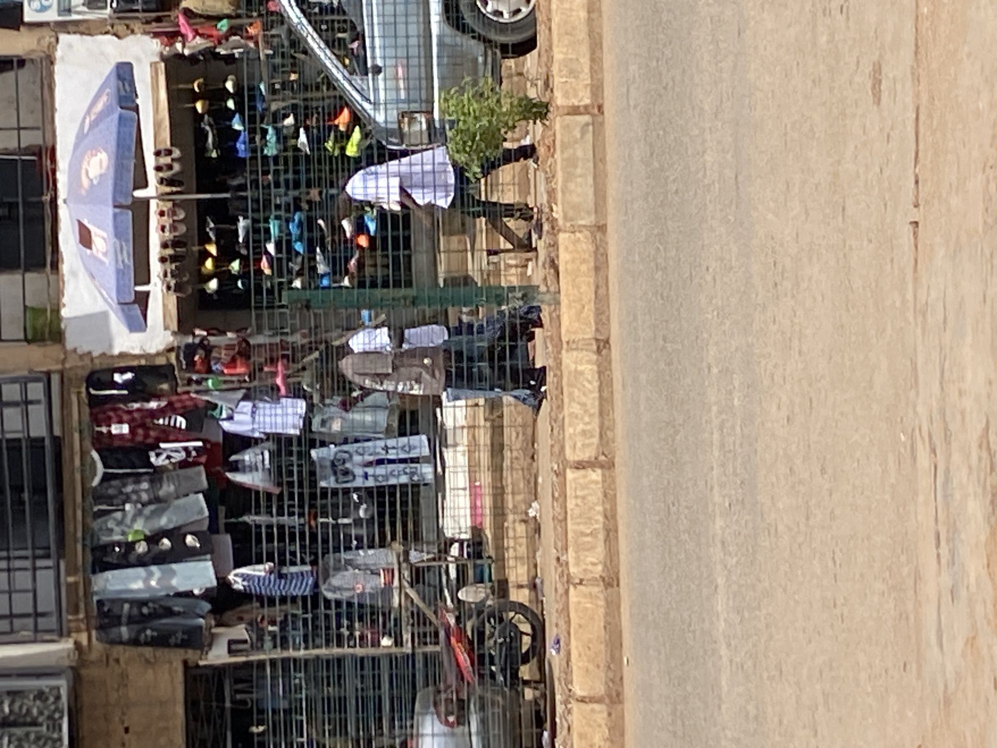 This image, captured in Bukuru, Nigeria, portrays an outdoor market scene during bright daylight. The perspective looks down onto a street with various stalls.
On the left, a vibrant street market is set up, featuring numerous metal racks and shelves displaying a wide array of merchandise. Clothing items like shirts (including plaid patterns, solid colors, and graphic tees), trousers, and shoes are prominently arranged for sale. One particular dark shirt on a rack shows what appears to be the "NIKE" logo. Another white shirt has "S B" and "89" printed on it. A large blue and white umbrella, likely providing shade for a stall, is visible at the top left, with an indistinct logo or branding on its surface. Beneath the umbrella, rows of shoes are displayed on shelves.
At least two individuals are visible within the market area. One person, wearing a white shirt and dark pants, is partially obscured by the metal fencing of a stall. Another figure in dark attire can be seen amidst the clothing displays further back.
Adjacent to the market stalls, a light-colored paved road occupies the right side of the frame, separated by a light brown stone curb. A silver sedan is parked along the curb, with its front wheel and a portion of its body visible behind the market setup. A small potted plant sits on the ground near the car. Closer to the bottom center, part of a bicycle or motorcycle wheel is visible. The scene suggests a bustling commercial area, indicative of daily life in a Nigerian urban setting.