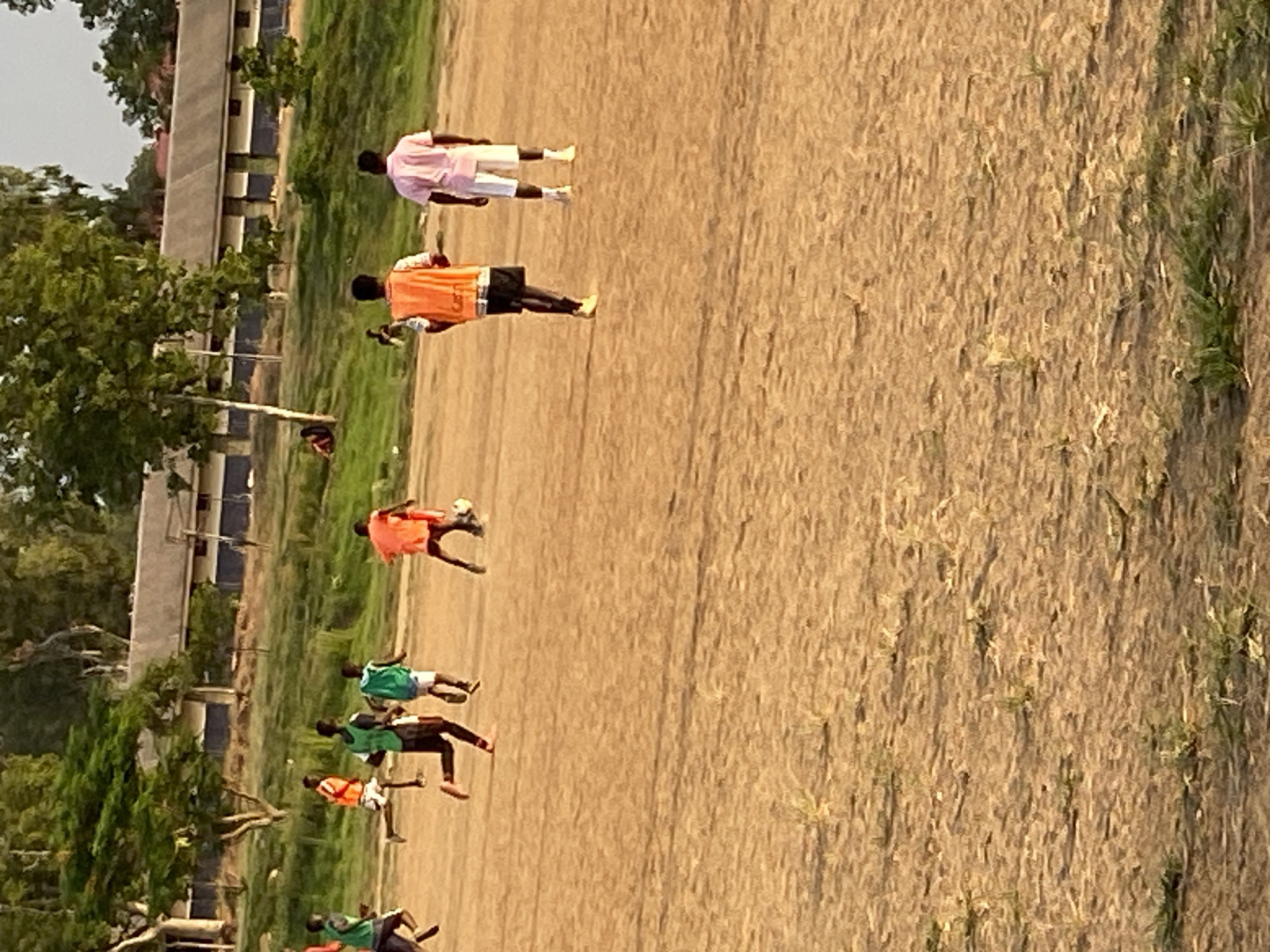 The image depicts a group of people playing soccer on a dirt field in Bukuru, Nigeria. Seven individuals are visible, wearing a mix of orange, green, and pink athletic shirts. Most are in motion, kicking or running. The field is dry and dusty, with some patches of green vegetation. In the background, there's a building with a red roof and trees lining the perimeter of the field. The scene appears to be in daylight with clear skies.