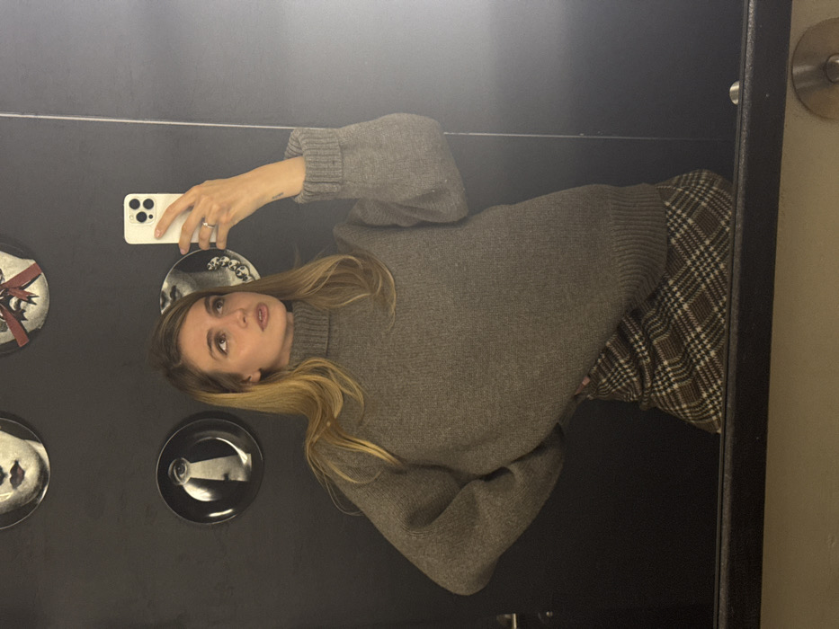 A woman with long, blonde hair wears a gray, oversized sweater and a plaid skirt. She holds a white iPhone in her right hand, taking a selfie in front of a mirror. The background includes a black wall decorated with several white-and-black plates featuring different illustrations. The overall tone is casual and trendy.