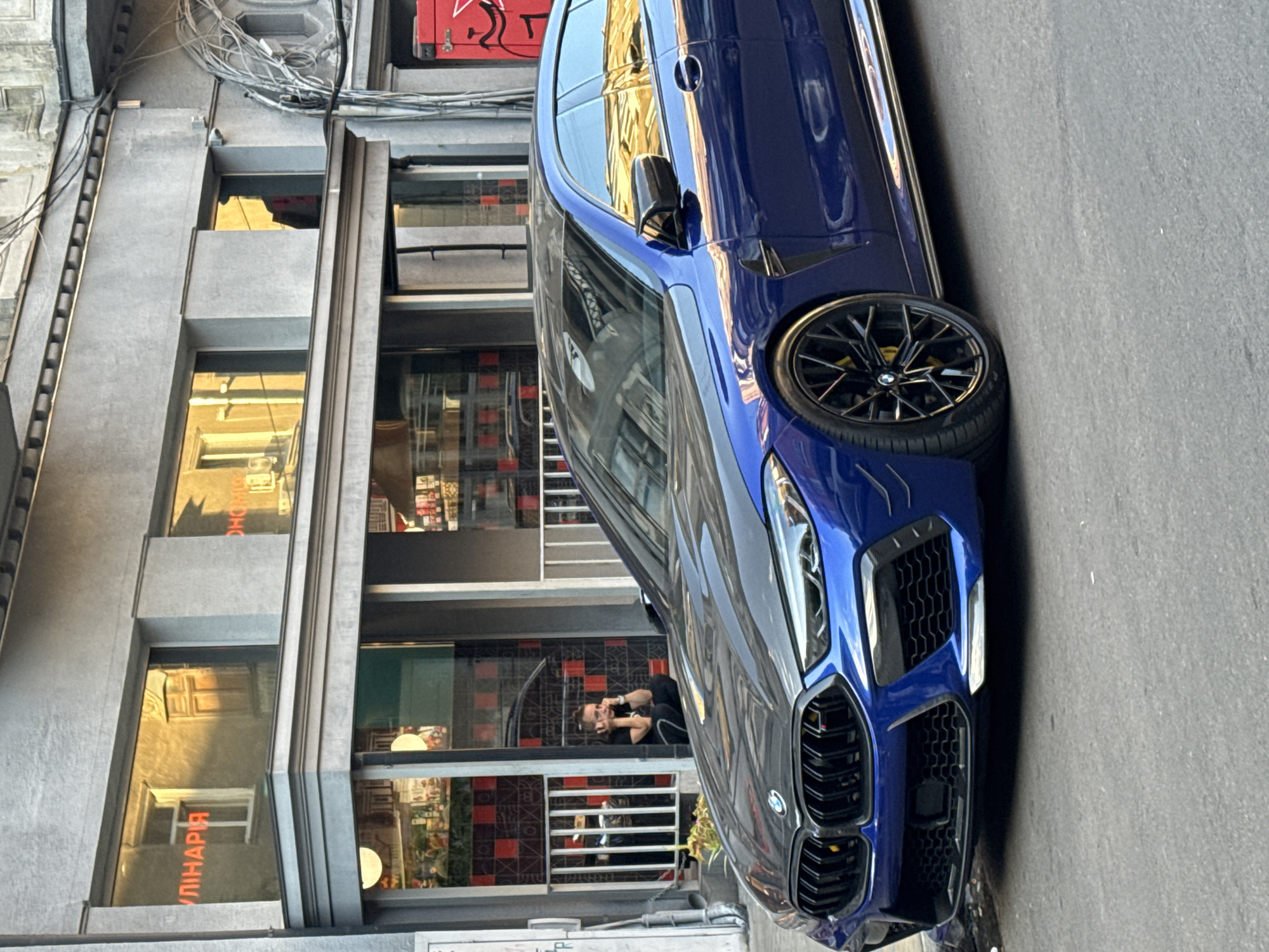 A blue BMW M8 Competition Gran Coupe is parked on a paved street in front of a building with a grey facade. The building features large windows with reflections of golden sunlight. Red neon signage on the building reads "КУЛІНАРІЯ" (Kulynariya, meaning "Culinary") in one window and appears to say "ОНОВЛЕНО" (Onovleno, meaning "Renewed") in another. A person is crouched in front of a white railing on the sidewalk, leaning their head on their hands. The car has a carbon fiber hood and black rims with yellow brake calipers. The overall scene suggests a commercial street in Odesa, Ukraine, with a focus on the car and the architectural elements of the building.