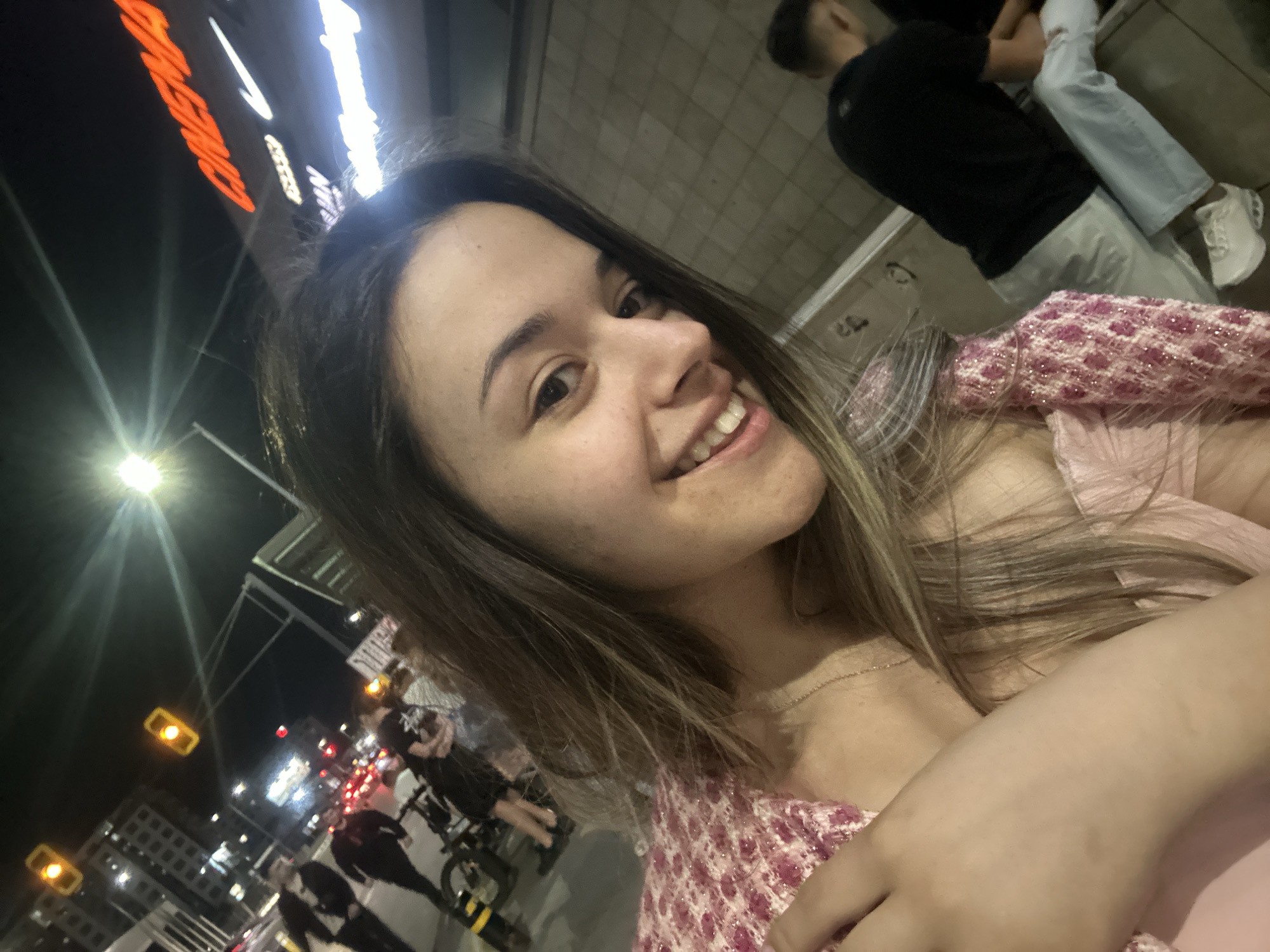 The image depicts a nighttime urban scene, with a young woman in the foreground smiling and looking towards the camera. She has long brown hair with lighter highlights, a light pink top, and a textured, sparkly pink overlay or cardigan. A thin gold chain is visible around her neck.
The background on the left reveals a street with blurred pedestrian traffic and distant vehicle taillights. Two illuminated yellow traffic lights are visible in the upper left, along with the dark silhouettes of urban buildings. A bright streetlamp emits a strong glare. A bicycle is partially visible near the bottom left, alongside a partially seen "STOP" sign.
On the right, a building facade features prominent illuminated signs. The word "CINEMA" in large orange letters is clearly visible, beneath which a white Nike swoosh logo is present. Further down, partial white illuminated text includes "AN" and other words. The lower section of this building exhibits a light brown/grey tiled or paneled texture. A male figure is seated on a low ledge or wall along the building's base, seen from behind, wearing a dark t-shirt, light trousers, and white sneakers. Another person's leg is partially visible beside him.
This combination of prominent "CINEMA" and Nike signage on a building, adjacent to a busy street with traffic lights and pedestrian activity, strongly indicates a commercial and entertainment district in Sofia, Bulgaria, likely associated with a major cinema complex such as Cinema City.