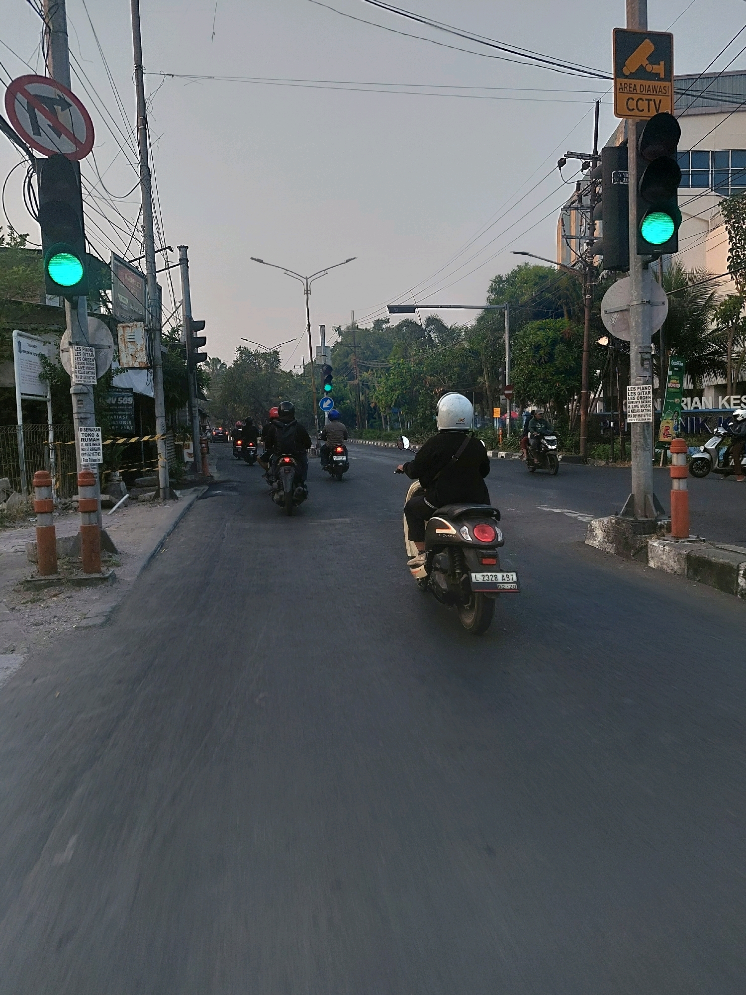 The image depicts a street scene in Surabaya, Indonesia, likely during daylight given the lighting conditions. The viewpoint is from a moving vehicle, indicated by the motion blur in the foreground and the angle of the shot. Several individuals on motorcycles are the primary subjects, navigating a paved road. The road appears to be a multi-lane street with some wear and tear, and a concrete barrier on the right side.
Traffic signals are visible, with at least one displaying a green light, suggesting traffic is permitted to proceed. A "No U-turn" sign is mounted on a pole on the left side of the road. A CCTV surveillance sign is also present on the right, indicating an "Area Diawasi CCTV" (Area Monitored by CCTV).
The environment includes a sidewalk on the left with some construction materials or debris, and a few orange traffic cones. On the right, beyond the concrete barrier, buildings and lush green trees are visible, suggesting an urban or suburban setting. Power lines crisscross overhead. The sky is overcast or hazy, typical of the urban atmosphere. The overall impression is a snapshot of daily life and traffic flow in a busy Indonesian city.