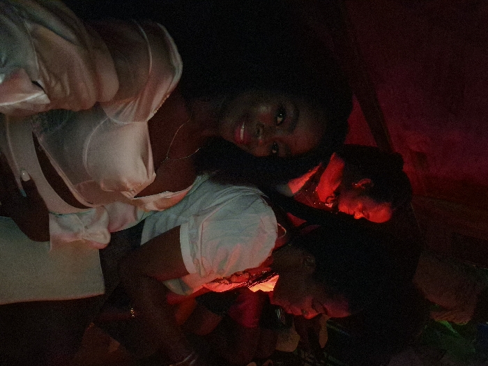 Three individuals are present in the photo. One person is visible from the chest up and is dressed in white, wearing a necklace. The other two people are visible from the neck up and the image appears to be indoors in a setting that is dimly lit with red light and appears to be in the dark.