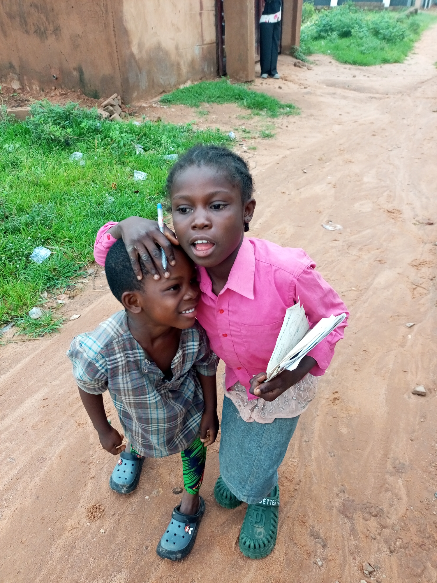 In Gombe, Nigeria, two children are captured in a portrait. The older child, a girl with dark skin and braided hair, wears a vibrant pink collared shirt and a light-colored skirt. She holds a stack of papers in her right hand and a pen in her left, with her hand resting on the head of the younger child. The younger child, a boy with dark skin and short hair, is dressed in a plaid shirt and patterned leggings. He wears blue clogs with white charms. Both children are standing on a dirt path, with a grassy area and a building visible in the background. The overall scene suggests an outdoor setting, possibly in a residential area.