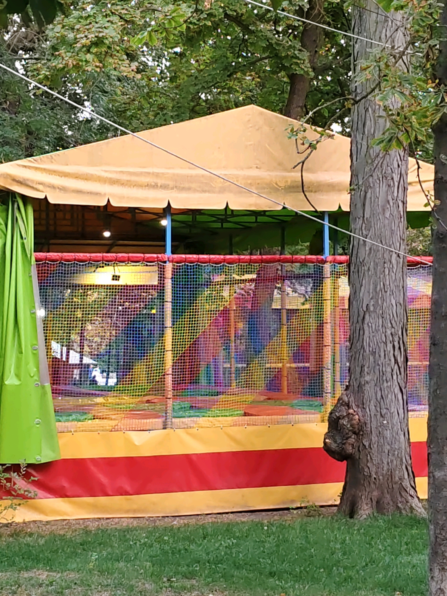 The image depicts an outdoor children's play area, likely a bounce house or soft play structure, enclosed by a yellow tent with a green tarp to the left. The structure features a colorful net enclosure with red trim and a base adorned with yellow and red stripes. Inside the play area, various colorful slides and platforms are visible. The scene is set amidst a backdrop of lush green trees, and a large tree trunk with a prominent burl is situated in the foreground to the right, partially obscuring the play structure. The ground is covered in green grass. The image appears to have been taken in the city of Rostov-on-Don, Russia, possibly in a park or recreational area. There are no people clearly visible within the play structure, but the overall impression is of a child-friendly outdoor entertainment venue.