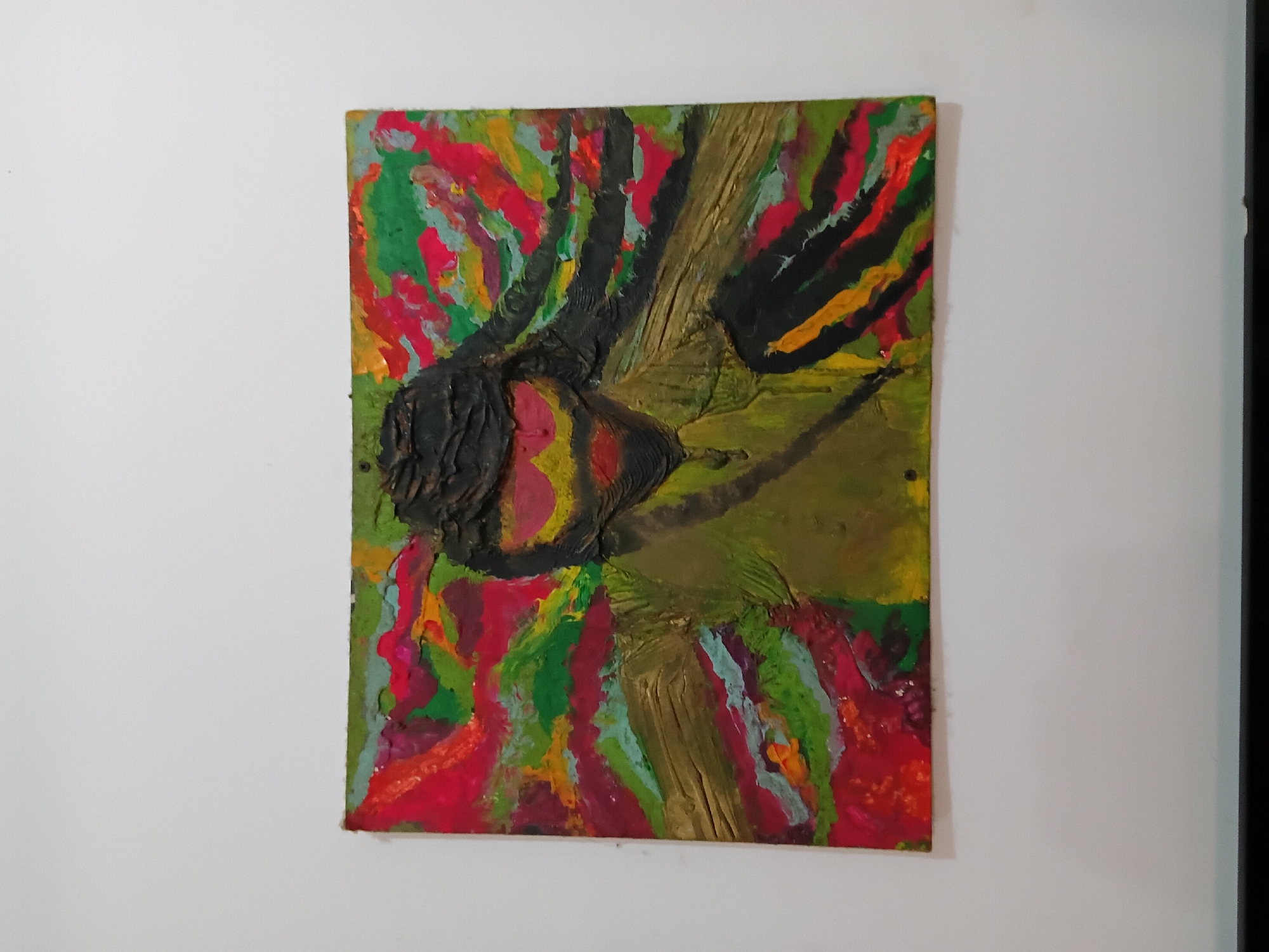 A painting is mounted on a white wall. The painting features thick, textured strokes of color. Dominant colors include red, green, yellow, brown, and black. The overall impression is abstract and somewhat crude, with no identifiable subject matter clearly defined. The location is Chopdem, India.