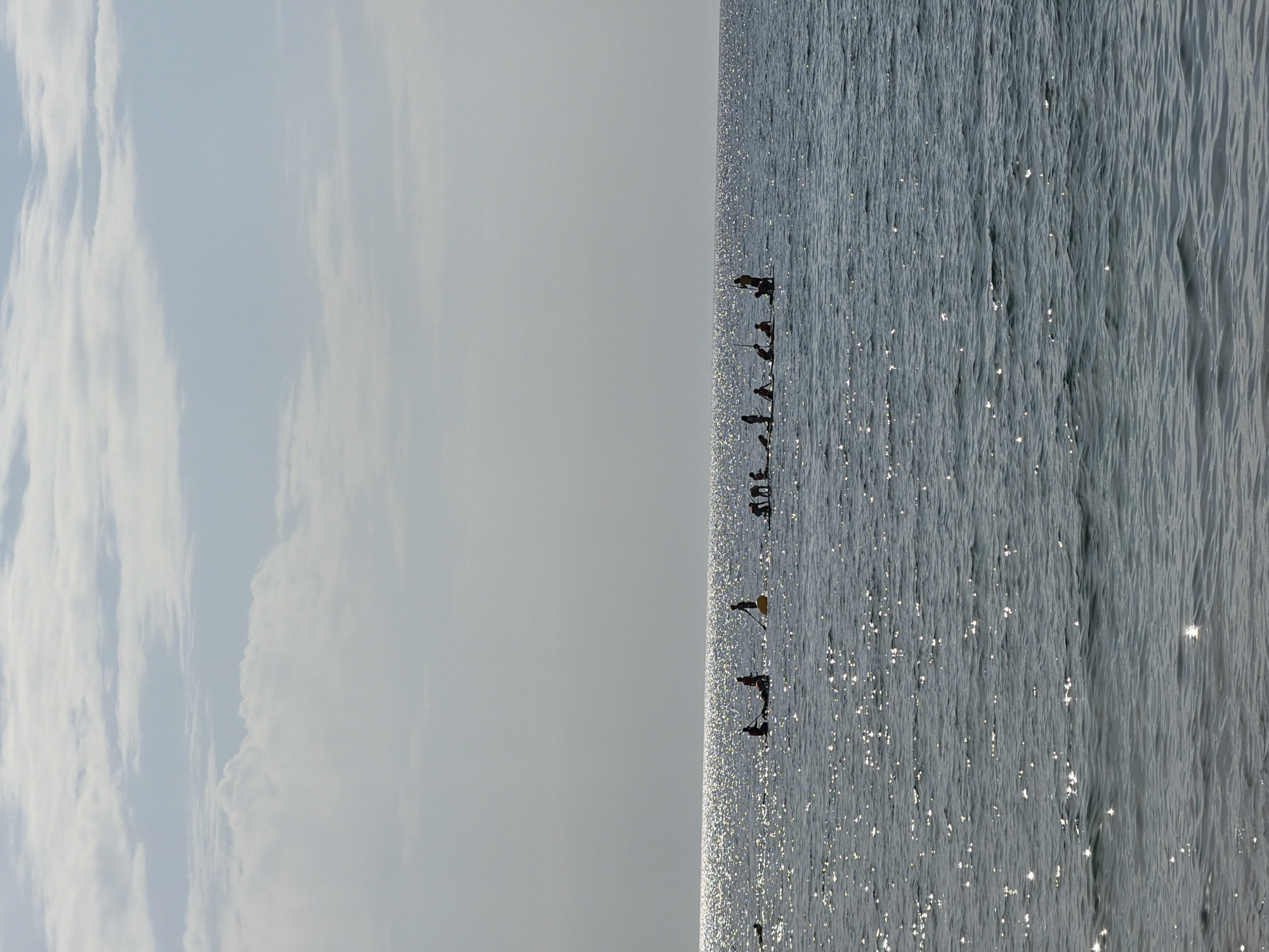 In Barcelona, Spain, this image captures a serene ocean scene under a hazy sky with scattered clouds. The sun glints off the water's surface, creating a shimmering effect. A group of people are engaged in paddleboarding and kayaking in the distance. Some are standing on their boards, while others are seated. They appear to be participating in a group activity, possibly a lesson or a tour. The water is calm with gentle waves. The overall atmosphere is peaceful and recreational.