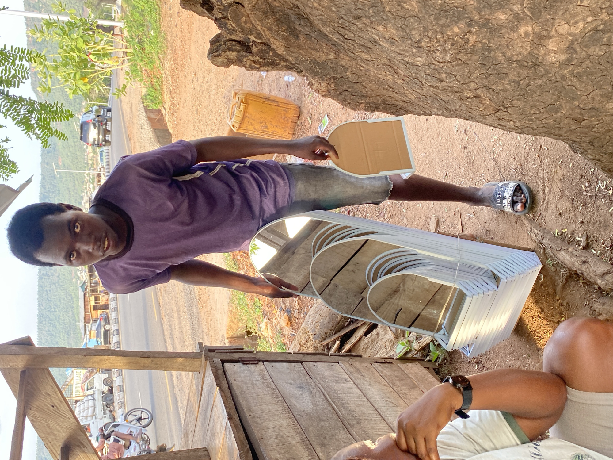 This image, rotated 90 degrees counter-clockwise, captures an outdoor scene at a roadside location, likely a market or stall area in Adankolo, Nigeria.
The main subject is a young Black man standing towards the left of the frame. He wears a purple short-sleeved t-shirt, faded grey shorts, and black flip-flops with white text that reads "LUOFU". He looks directly at the camera with wide, curious eyes and a slightly open mouth. In his right hand, he holds a light brown, rounded rectangular piece of cardboard. To his immediate left, a stack of white-framed, oval-shaped mirrors rests on the ground. Behind him, a yellow plastic jerrycan is visible on the dirt.
In the bottom right corner, a second individual, likely a man, is partially visible, sitting. Only his torso, right arm, and legs are seen. He wears a light-colored t-shirt and shorts, with a dark-strapped watch on his left wrist, and appears to be looking downwards.
The background features a paved two-lane road with a white center line, indicating traffic flow. Several vehicles are present, including a dark SUV, other cars, a white truck laden with goods that displays the text "TOMITI VILLAGE," and at least one motorcycle. Beyond the road, green hills and lush vegetation stretch into the distance under a bright, clear sky, suggesting either late morning or afternoon. Large tree trunks and wooden structures, possibly parts of stalls or shelters, frame the scene. The ground is a mix of dirt and dry foliage. The overall impression is a bustling yet typical roadside environment.