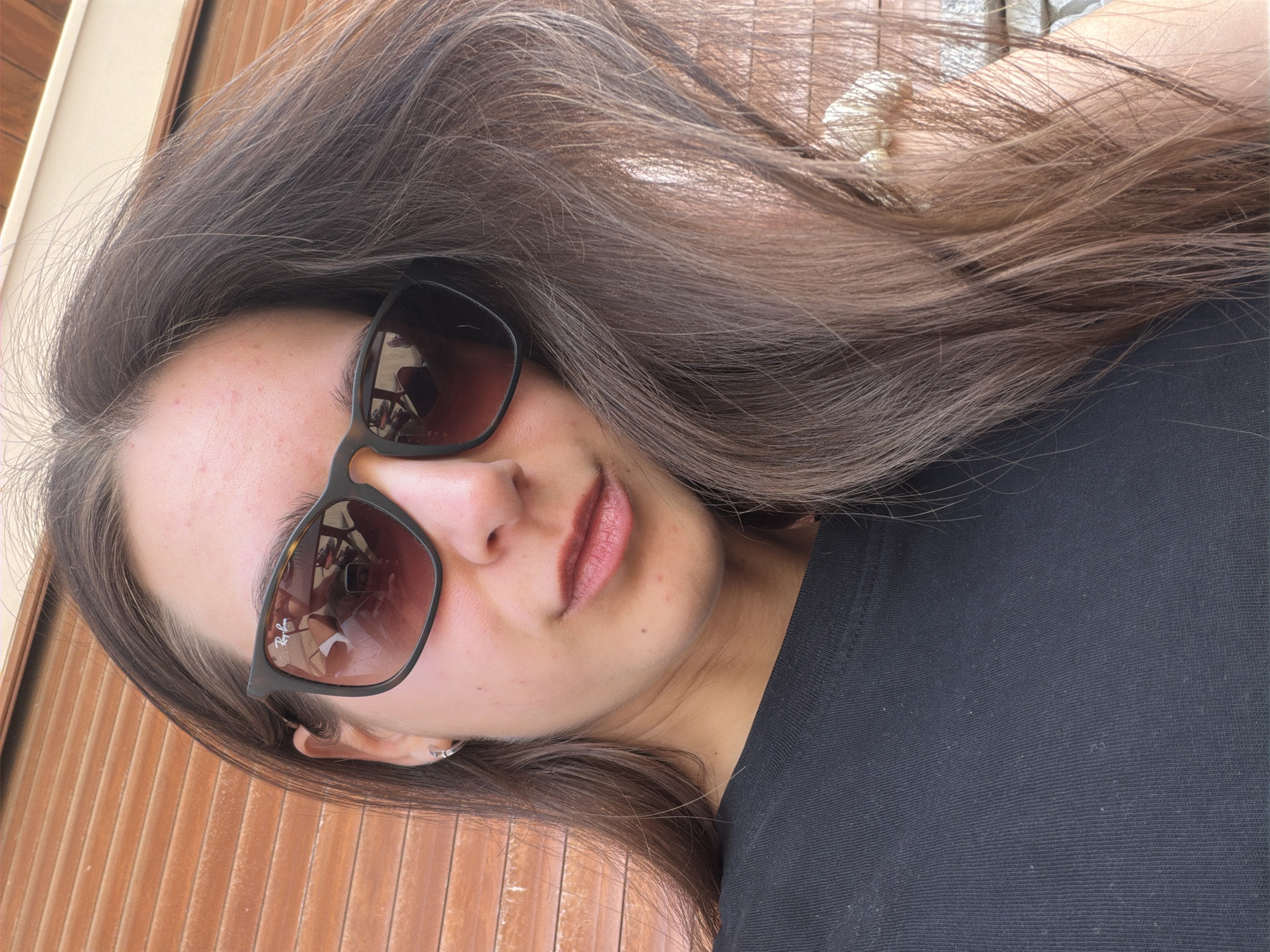 A young woman with long brown hair and wearing dark sunglasses is posing for a selfie. The sunglasses reflect a beach scene. She is wearing a black t-shirt. The background is a wooden door or wall. The scene is likely in Boreti, Montenegro, based on the reflected beach scene.
