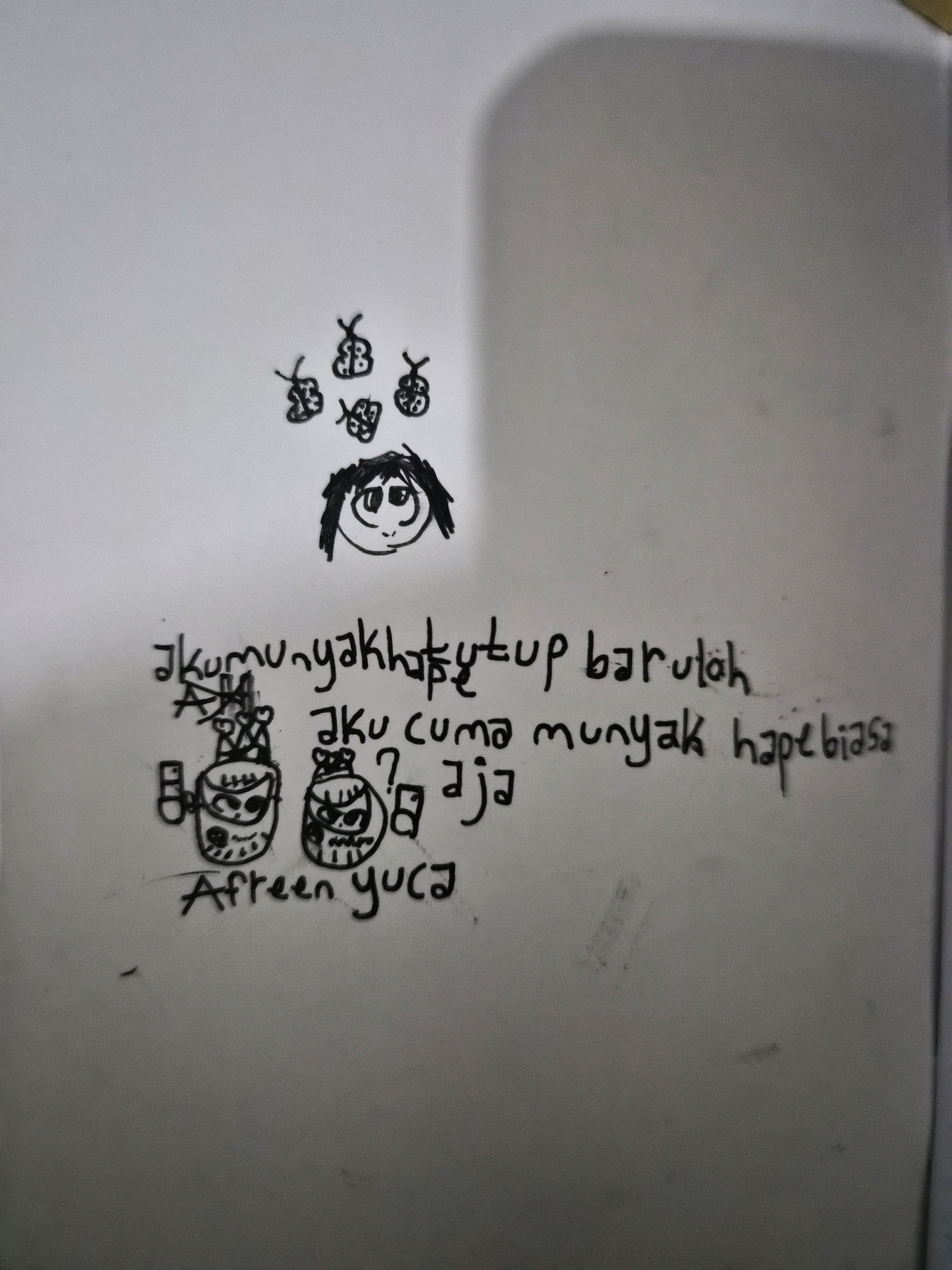 The image is a close-up, slightly angled, overhead shot of a doodle on a white surface, likely a whiteboard or paper, that appears to be located in Balikpapan, Indonesia. The doodle features several elements: a cartoonish face with large eyes and messy hair, surrounded by four small butterfly-like shapes. Below the face, there are two crudely drawn figures resembling robots or creatures with expressive faces, wearing crowns. To the right of these figures, there is handwritten text. The primary text reads "akumunyakhatutup barulalah". To the right of this, a smaller line of text says "aku cuma munyak hape biasa". Below the robot figures, the name "Afreen yu ca" is written. The overall impression is that of a whimsical, possibly child-like drawing with accompanying commentary. The background is largely out of focus, but the white surface dominates the frame.