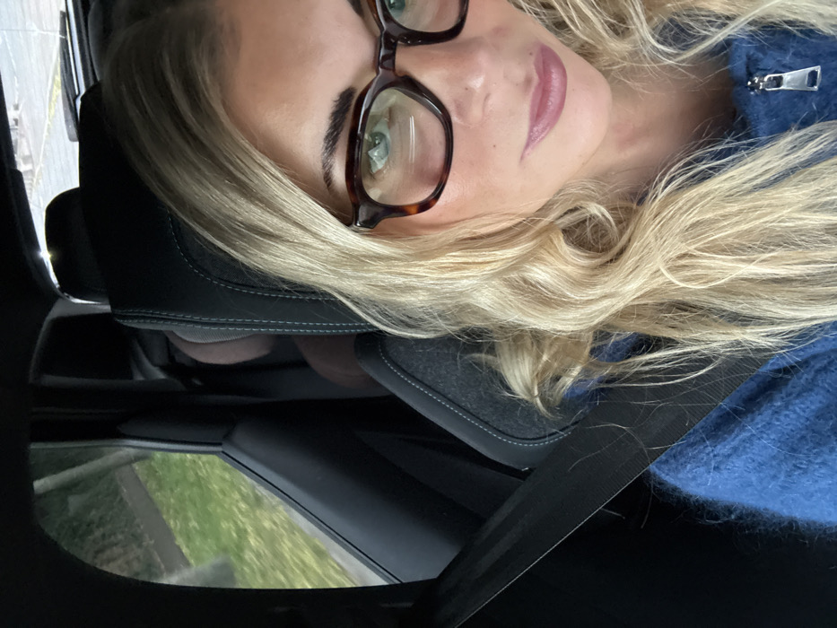An image captured in the city of Trets, France, depicts a female seated in the front passenger seat of a vehicle. She has blonde, wavy hair and is wearing dark, rectangular, tortoise-shell framed eyeglasses, revealing light-colored eyes. Her lips are a muted pink-mauve tone. She is dressed in a blue, fuzzy, zippered garment. A black seatbelt is visible across her chest and left shoulder. The car's interior features a dark gray fabric seat with light stitching, and dark interior trim. In the background, a dark brown padded headrest of another seat is visible. Through the passenger window, green foliage and what appears to be a light-colored ground or road surface are discernible, suggesting an outdoor environment.
