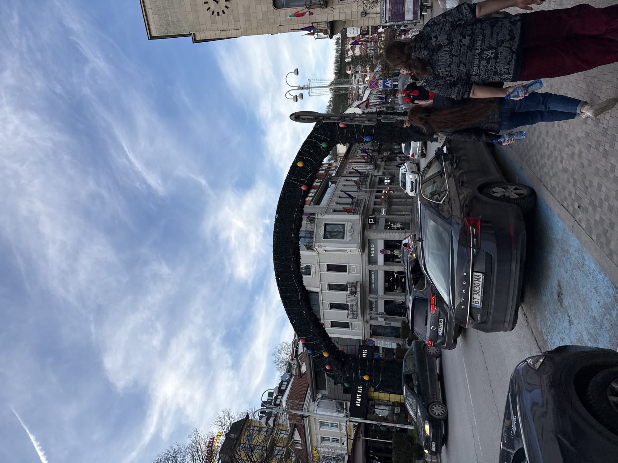 The image depicts a street scene in Veliko Tarnovo, Bulgaria. A prominent black arch, decorated with colored orbs and holiday greenery, spans the street, possibly for a seasonal event. The architecture along the street includes multi-story buildings, some with awnings and signage for stores such as Hugo, Karl Lagerfeld, and NESS. Several cars are parked along the road, with visible license plates reading "BT 3830 MA" and "BT 7171 MA." Two individuals are walking along the sidewalk, each holding a water bottle. A clock tower is visible in the background.