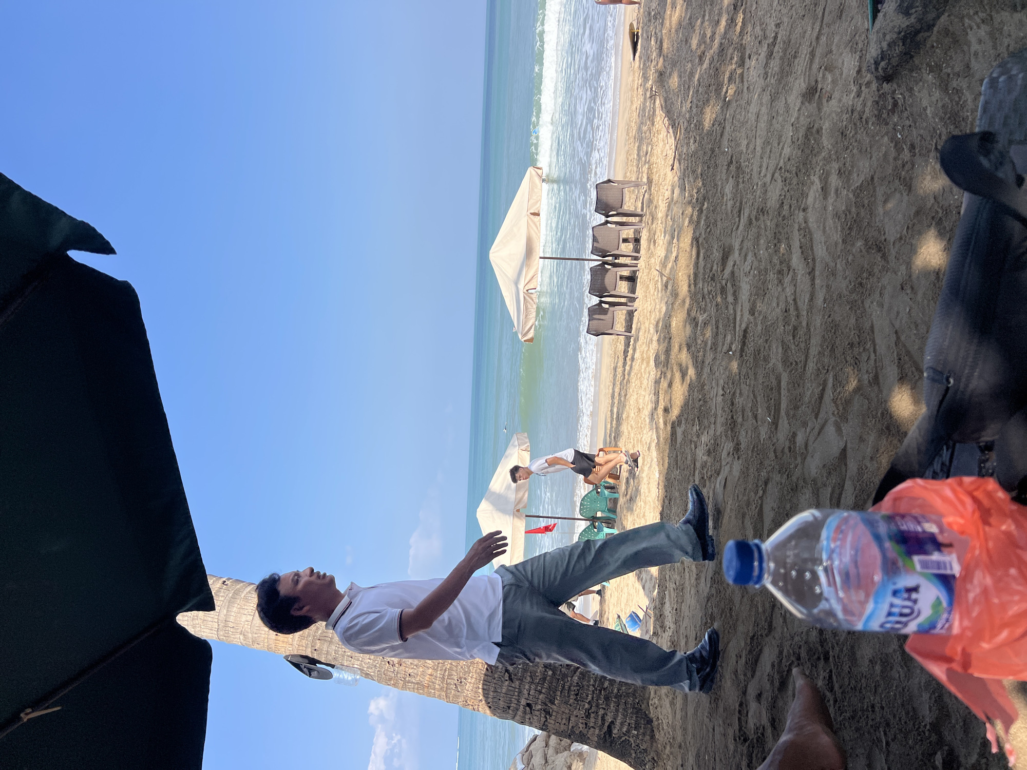 In Kuta, Indonesia, a man in a white polo shirt and gray pants walks across a sandy beach towards the ocean under a dark green umbrella. Palm trees and rocks are to his left. In the background, the blue ocean has moderate waves. Two beach umbrellas with chairs are set up further down the beach. Another man in a white t-shirt and black shorts is walking towards the water. A bottle of Aqua brand water is in the foreground on the sand, next to a red plastic bag and a black object. The sky is clear and blue.