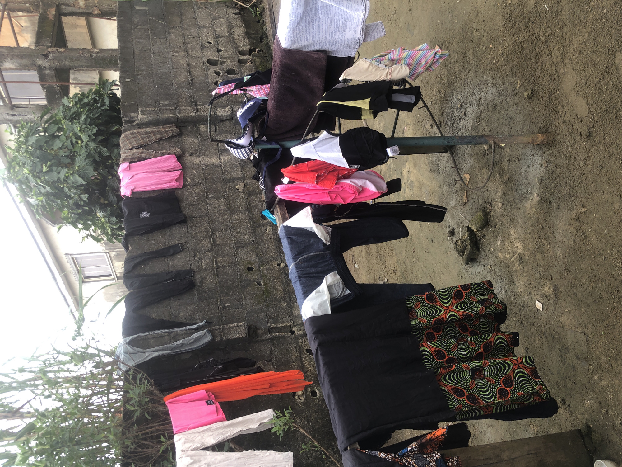 In a residential yard in Ado, Nigeria, laundry hangs on clotheslines and a drying rack in front of a rough-hewn concrete wall. The wall, appearing weathered and aged, has several circular holes, suggestive of construction damage or deliberate perforations. Behind the wall, a building with white siding and a single window can be seen, partially obscured by lush green foliage. The ground is a mixture of dirt and mud, with some scattered rocks and debris.
The laundry collection is diverse, featuring a variety of clothing items in different colors and patterns. On the upper clothesline, near the wall, several pairs of pants and shirts are hung, including a bright pink t-shirt, a pair of plaid pants, and black garments. Lower down, on a metal drying rack, a more colorful assortment is displayed: a black and orange patterned skirt, several brightly colored t-shirts (pink, red), white tops, and striped garments. A dark brown towel is also draped over the rack. The scene captures a common domestic activity, set against the backdrop of a typical urban Nigerian environment.